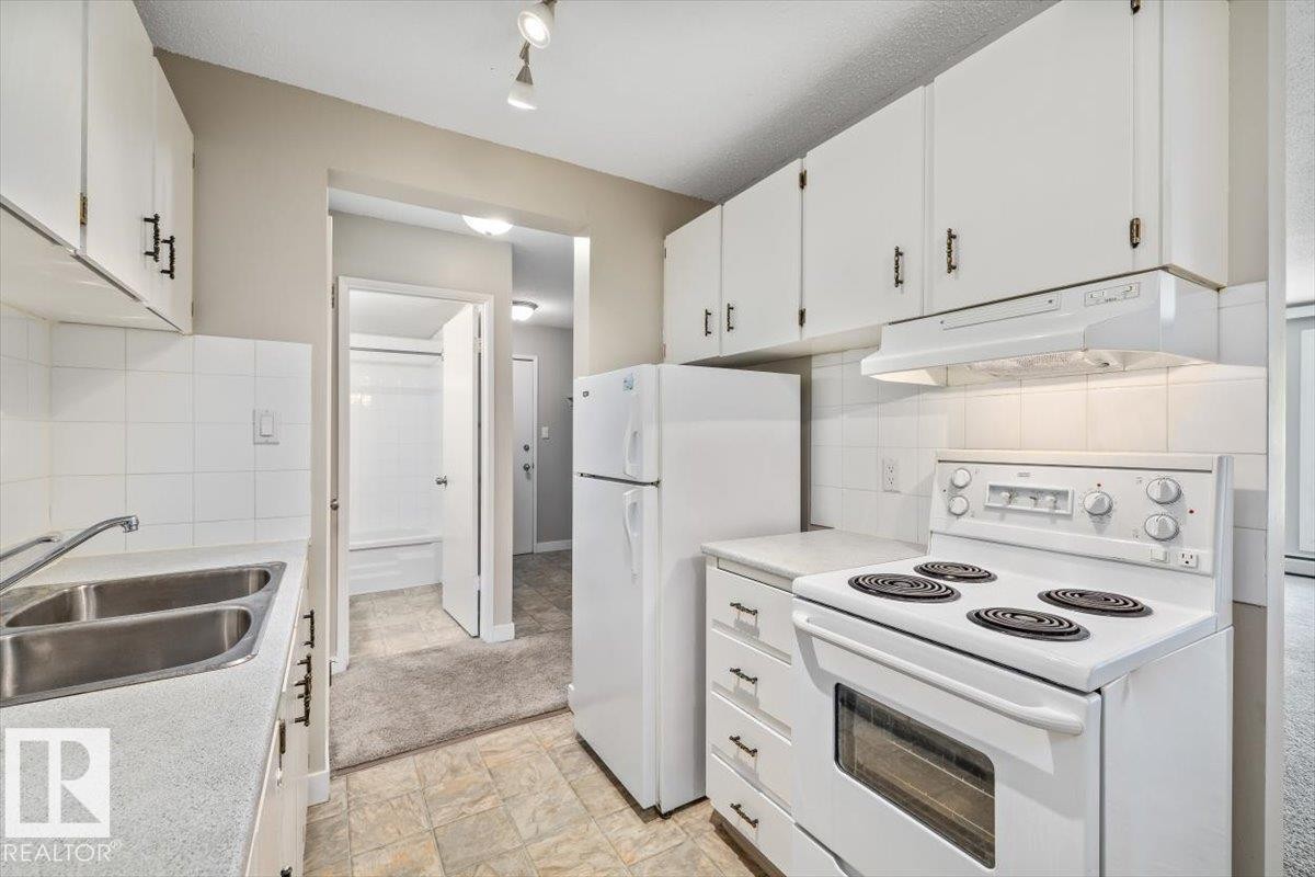 Kitchen featuring white upper and lower cabinetry, a double basin stainless steel sink, and a white electric range with overhead ventilation - #306 10330 113 St Nw, Edmonton, AB