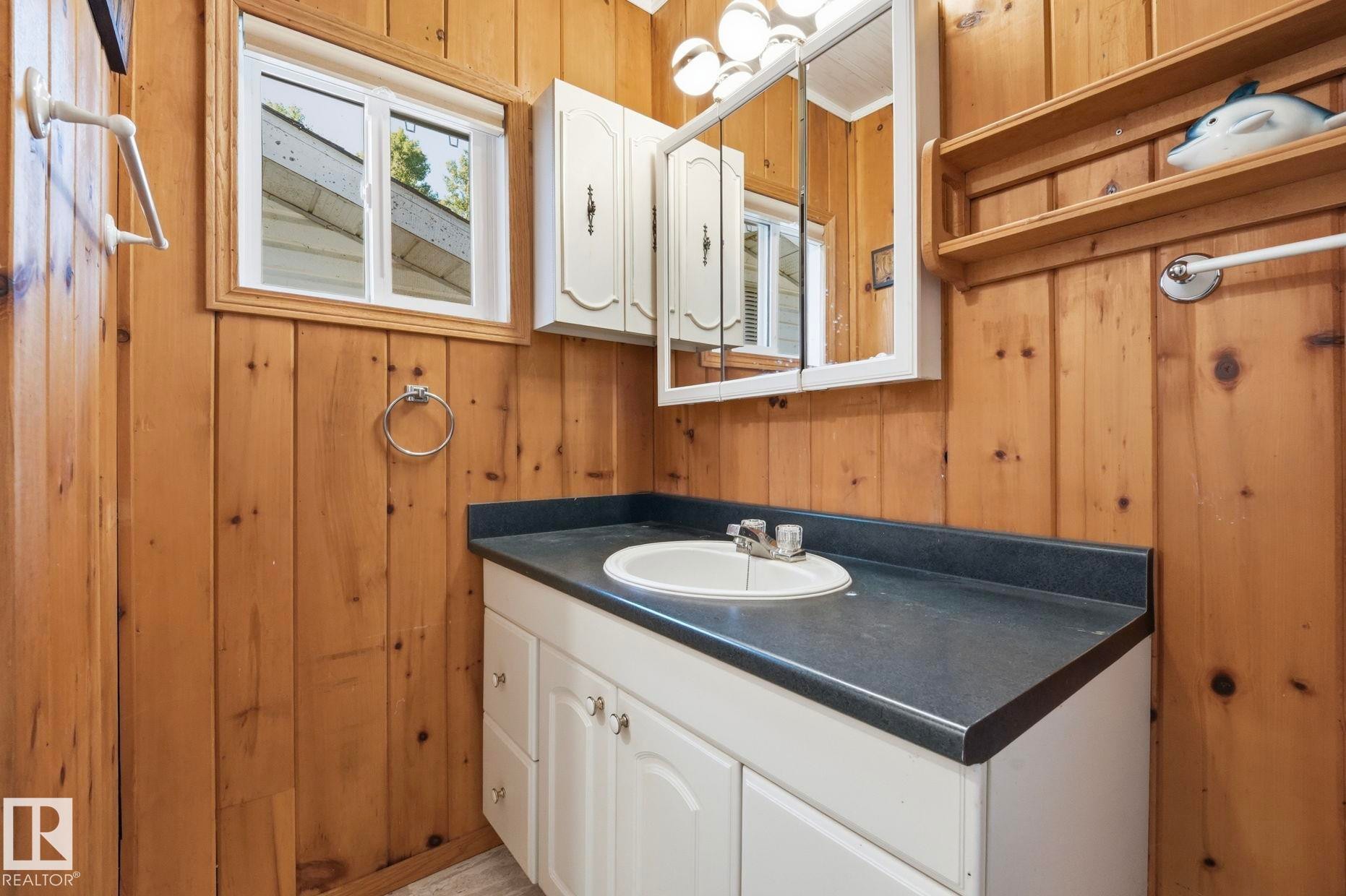 Bathroom featuring natural wood paneling, a white vanity with cabinet storage, a dark countertop with an integrated sink, a medicine cabinet with mirrored doors, and a window with a white frame - 1410 47436 Range Rd. 15, Rural Leduc County, AB