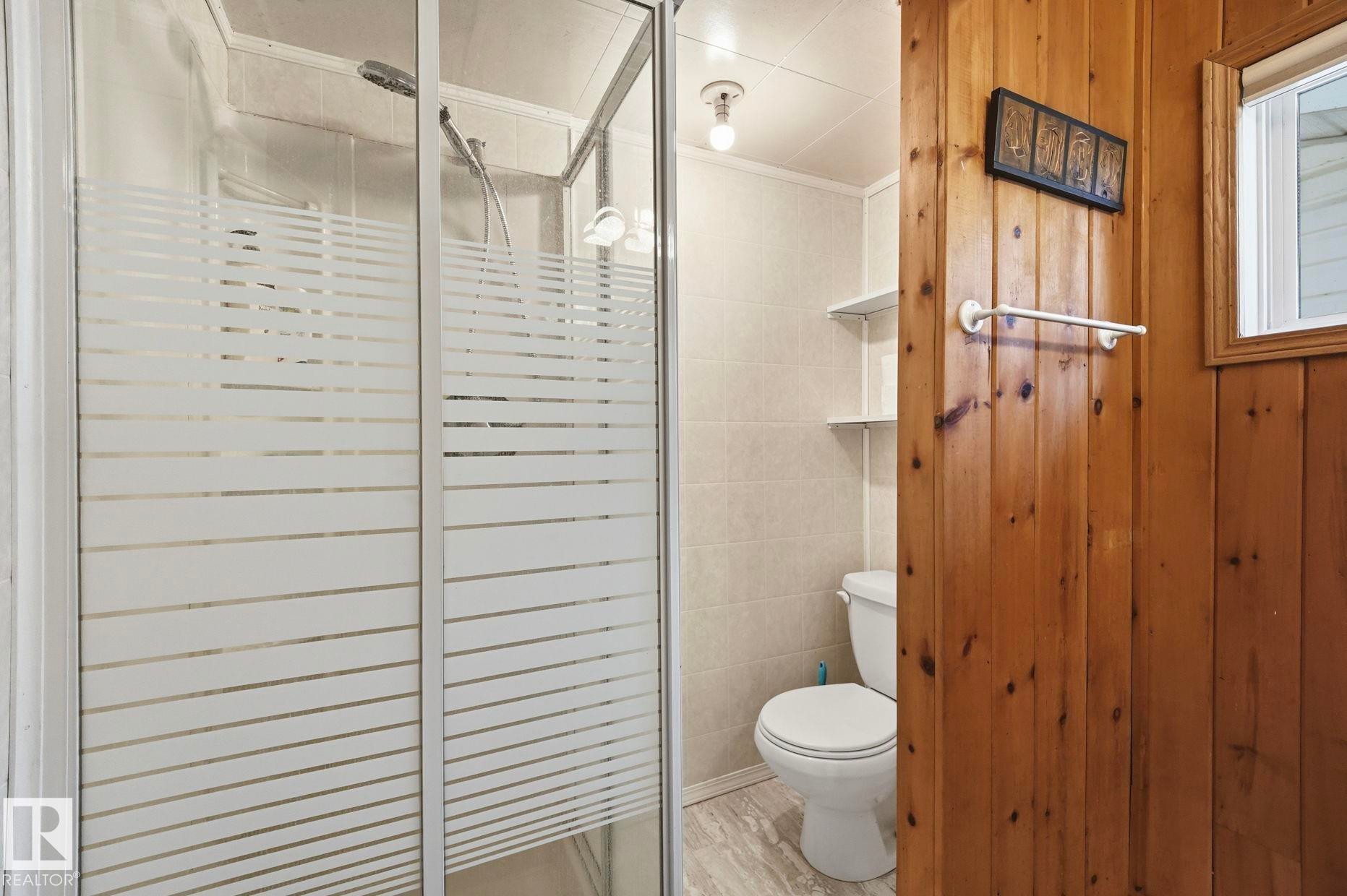 Shower enclosure with frosted glass doors, white ceramic tile wall, light-toned wood paneling, white toilet, and gray-toned floor tile - 1410 47436 Range Rd. 15, Rural Leduc County, AB