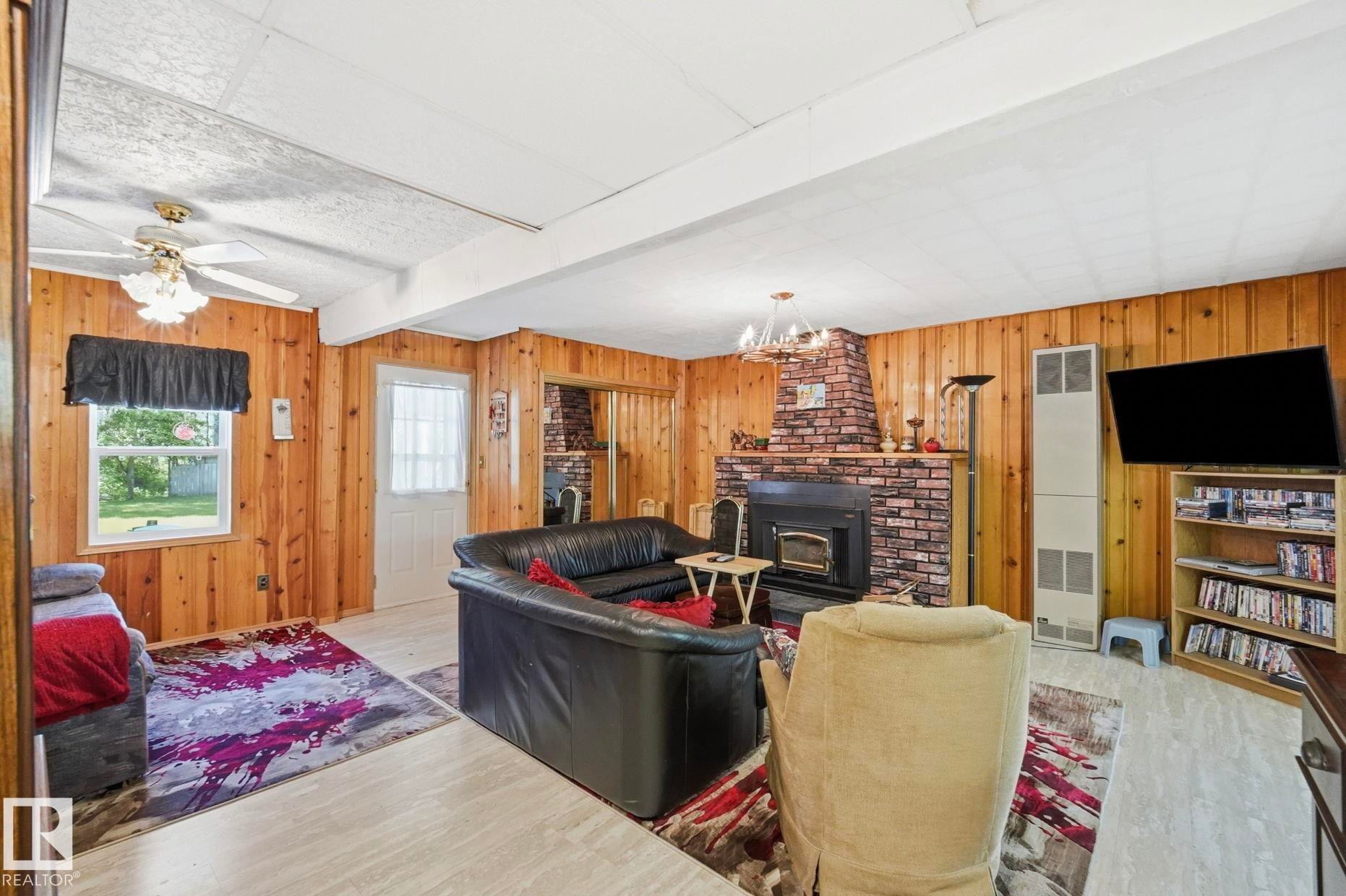 Living area featuring wood paneling, a brick fireplace, wood-finish flooring, a ceiling fan with light fixture, and a window with exterior views - 1410 47436 Range Rd. 15, Rural Leduc County, AB