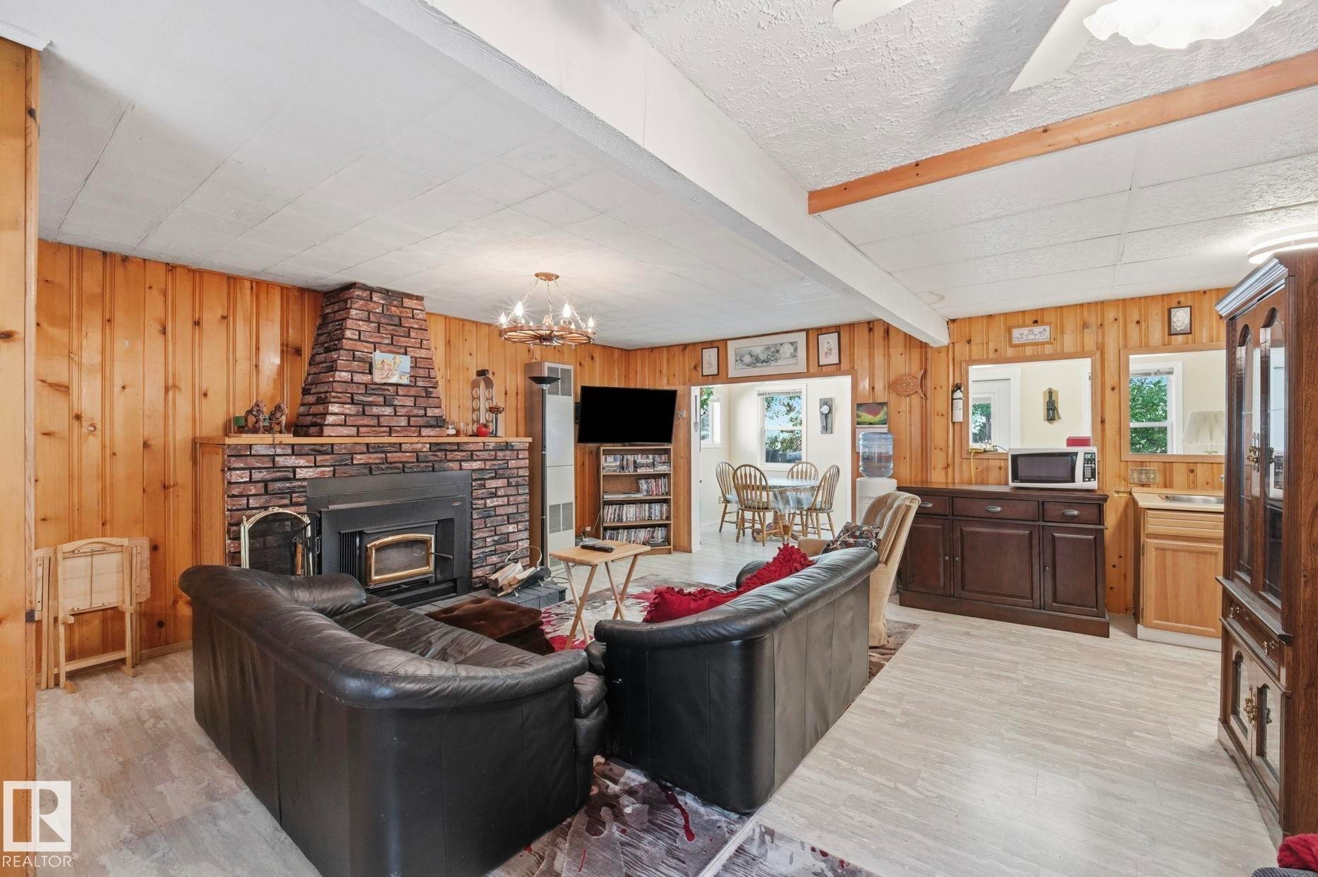 Living area featuring wood paneling, a brick-surround fireplace with an insert, light wood-finish flooring, and exposed ceiling beams - 1410 47436 Range Rd. 15, Rural Leduc County, AB