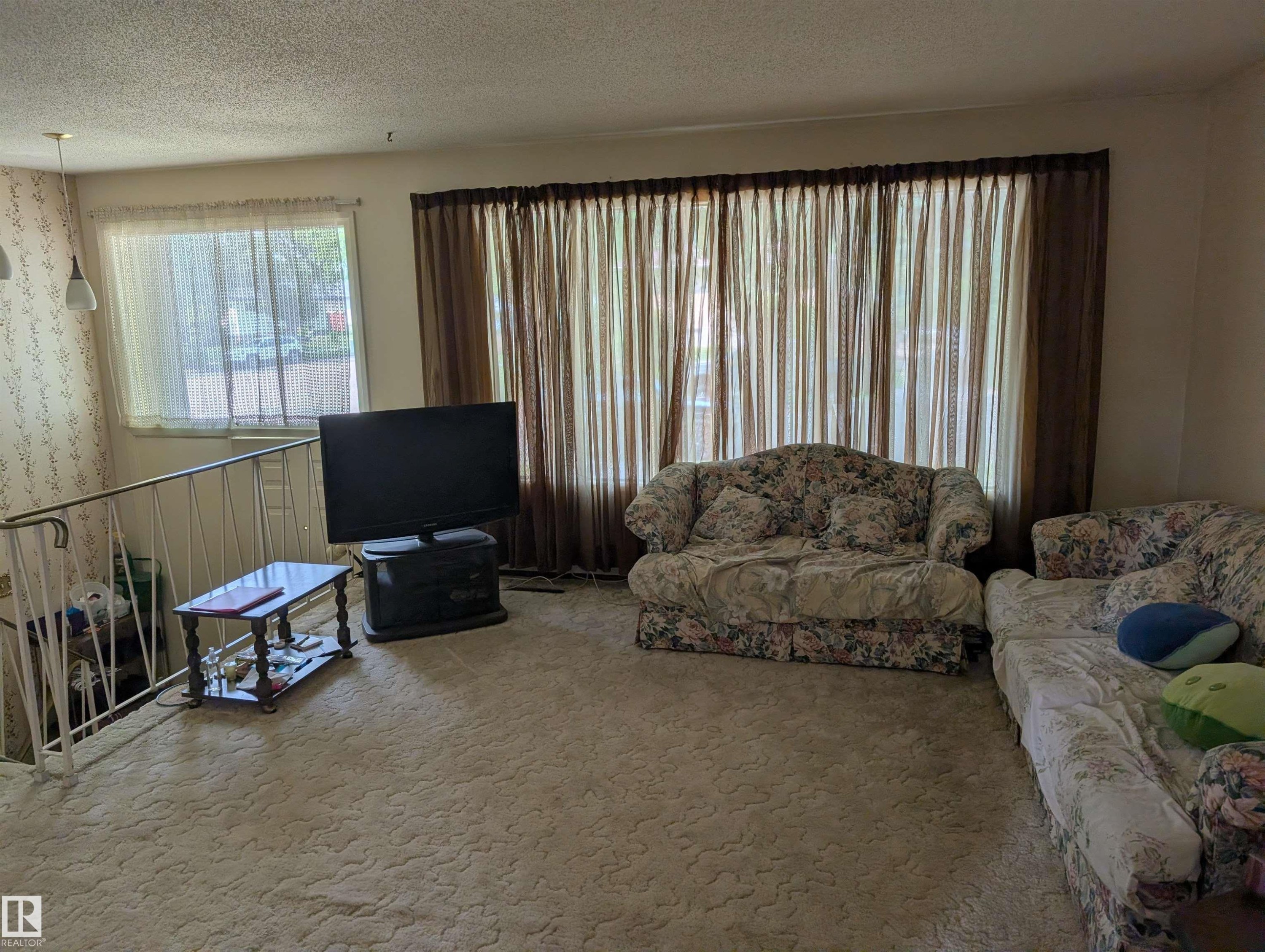 Living area featuring a large window with sheer and blackout curtains, carpeted flooring, and a white metal railing - 6415 147 Avenue, Edmonton, AB