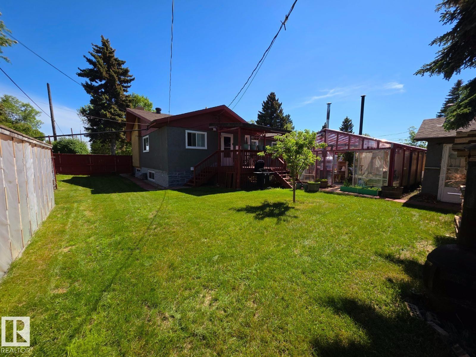 Expansive backyard featuring a lush lawn, a multi-paneled greenhouse, a raised wood deck with stairs, mature perimeter trees, and a privacy fence - 10404 116 Avenue, Edmonton, AB