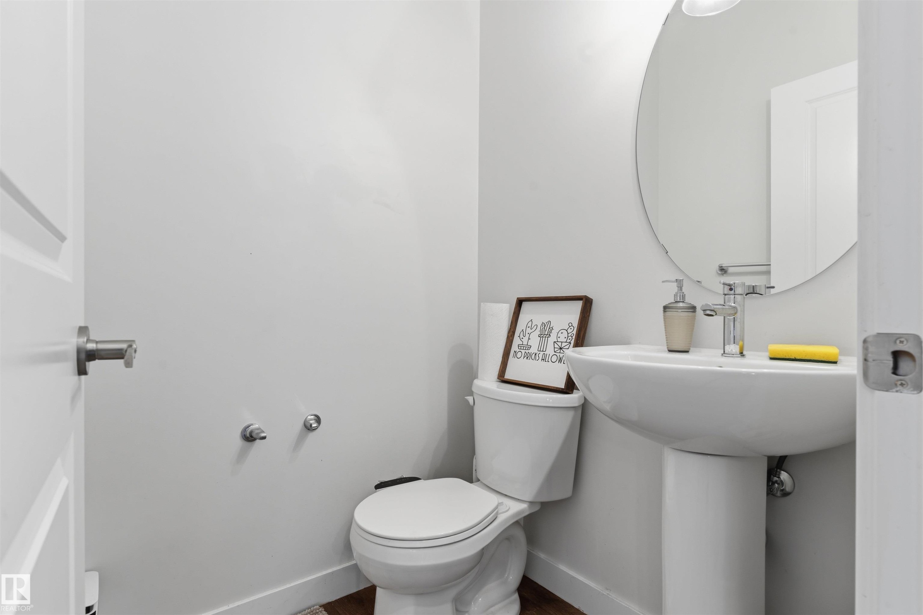 Contemporary powder room featuring a pedestal sink with chrome faucet, a white toilet, a round wall-mounted mirror, and wood-finish flooring - 170 804 Welsh Drive, Edmonton, AB
