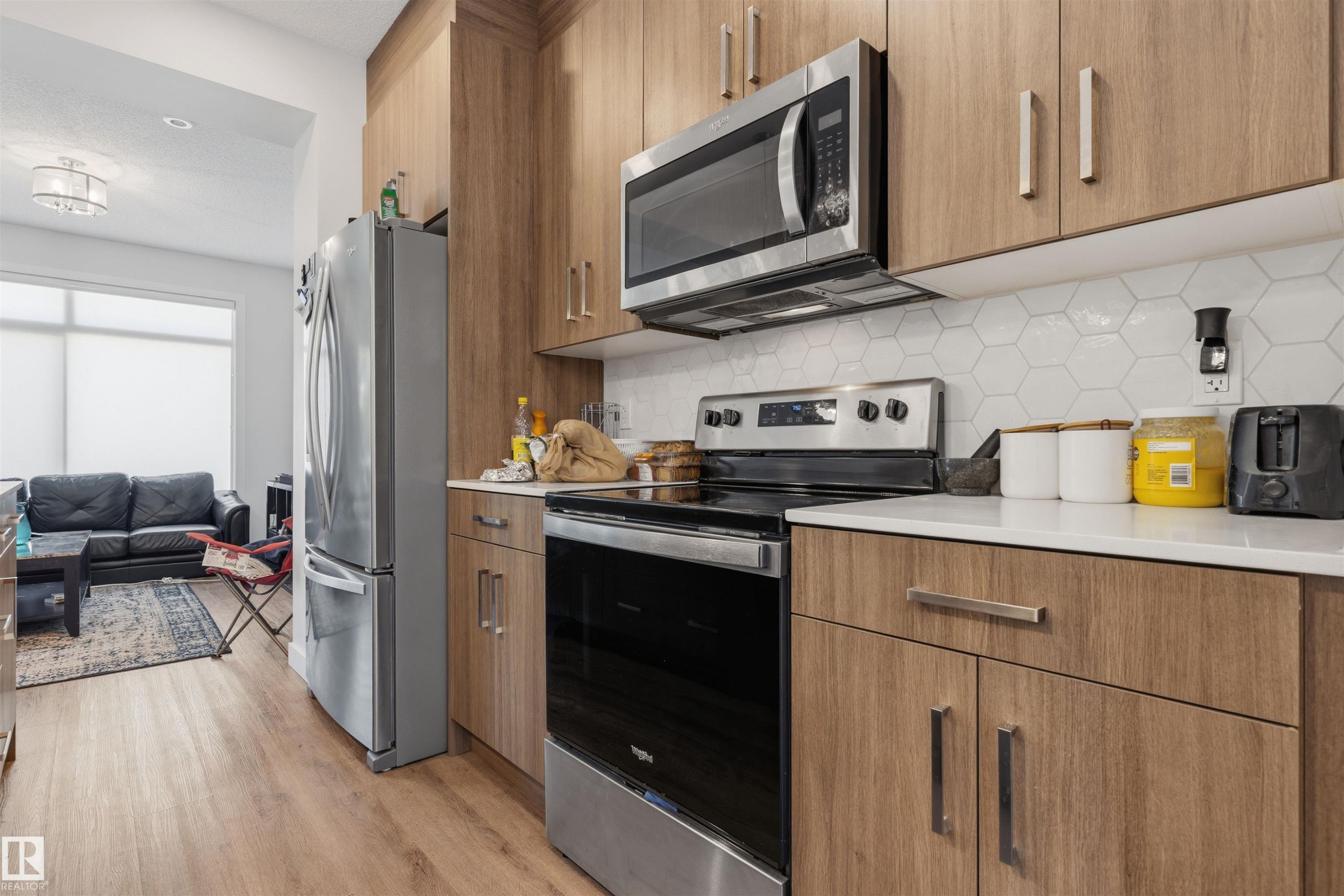 Modern kitchen featuring wood-finish cabinetry, stainless steel appliances, white hexagon tile backsplash, and light-toned countertops - 170 804 Welsh Drive, Edmonton, AB