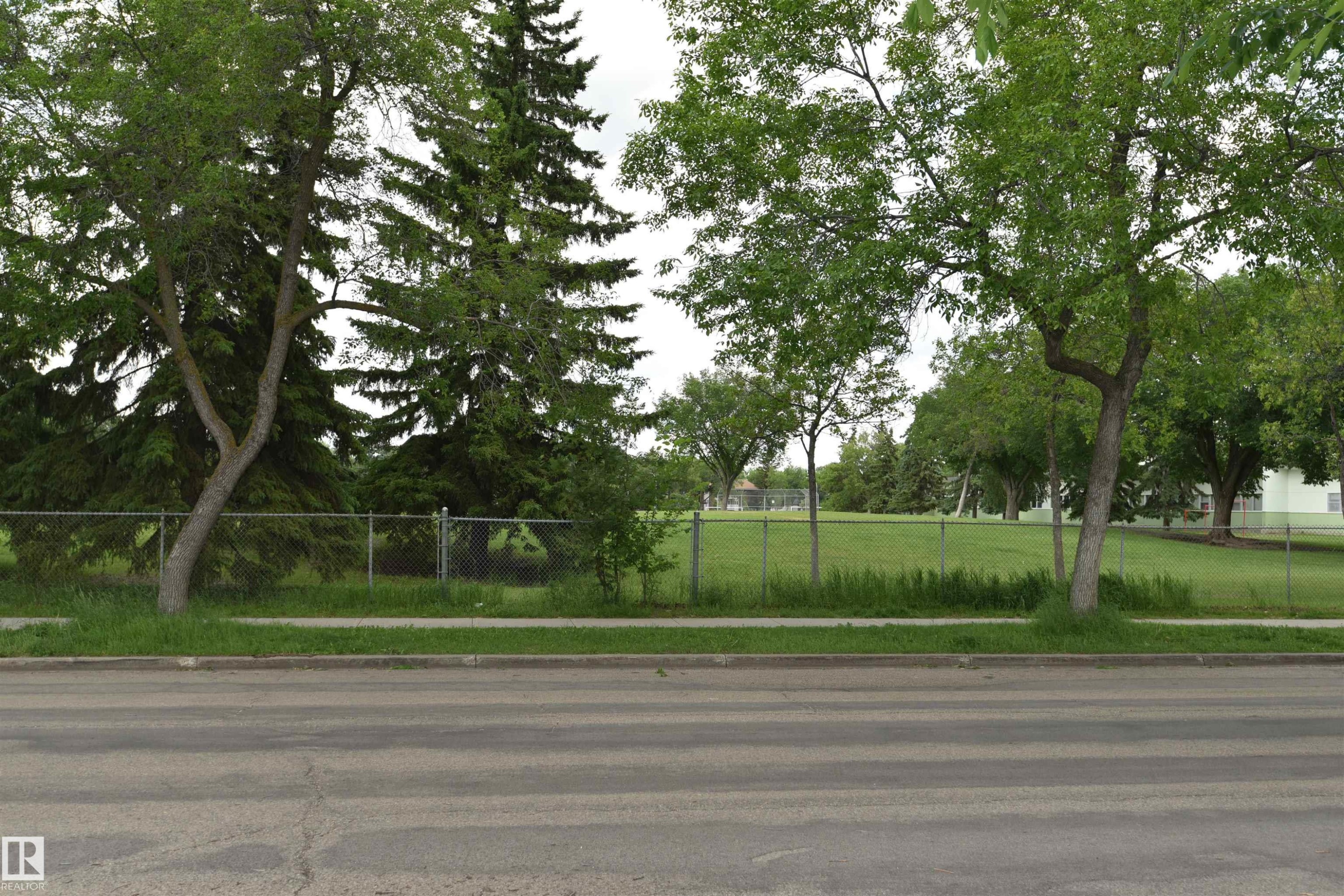 Expansive green space featuring a chain-link fence, mature trees, and a paved sidewalk along a street - 7923 94 Avenue Nw, Edmonton, AB - Outdoor