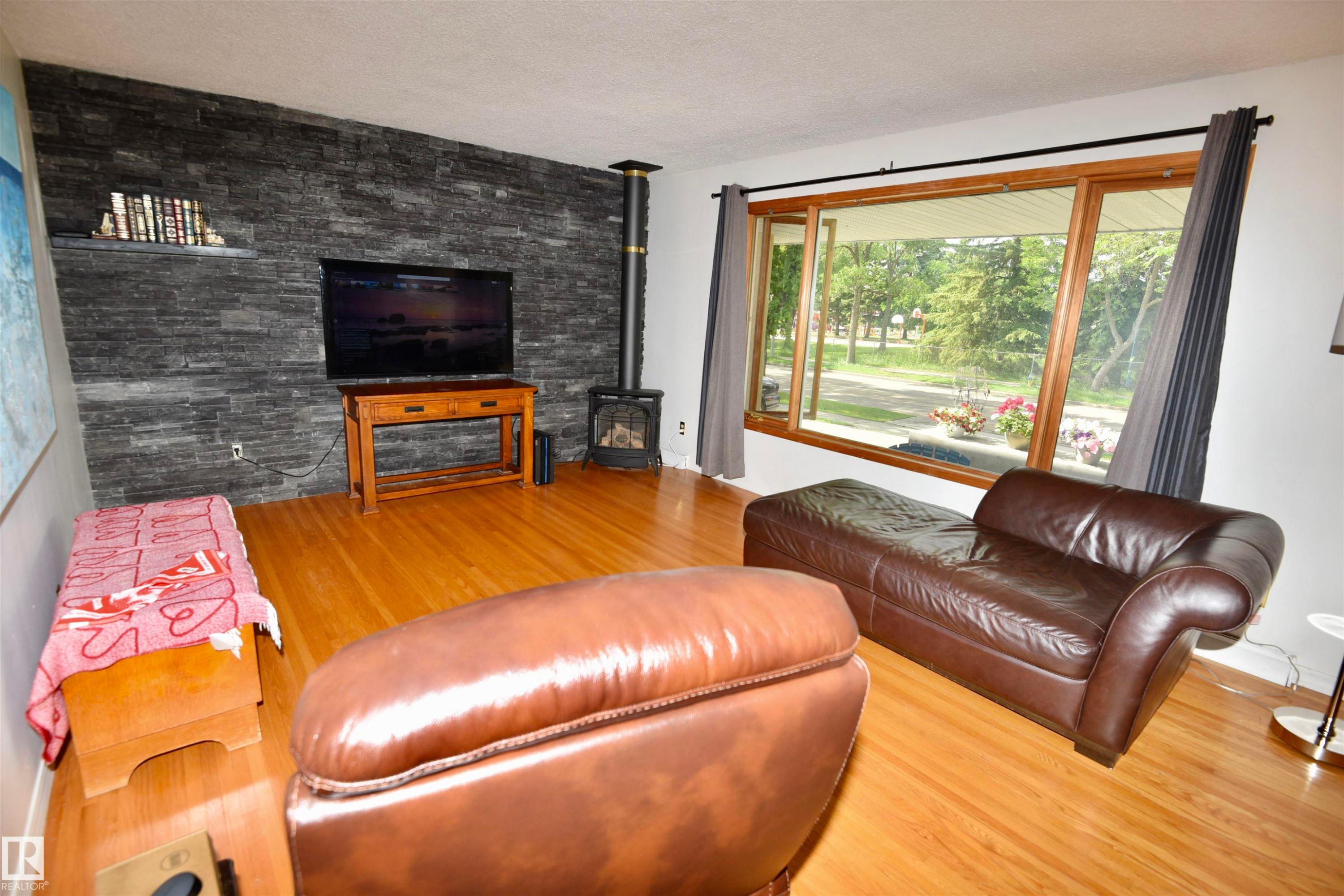 Living area featuring wood-finish flooring, a wood-burning stove, a large picture window, and a stone accent wall - 7923 94 Avenue Nw, Edmonton, AB - Indoor Photo Showing Living Room With Fireplace