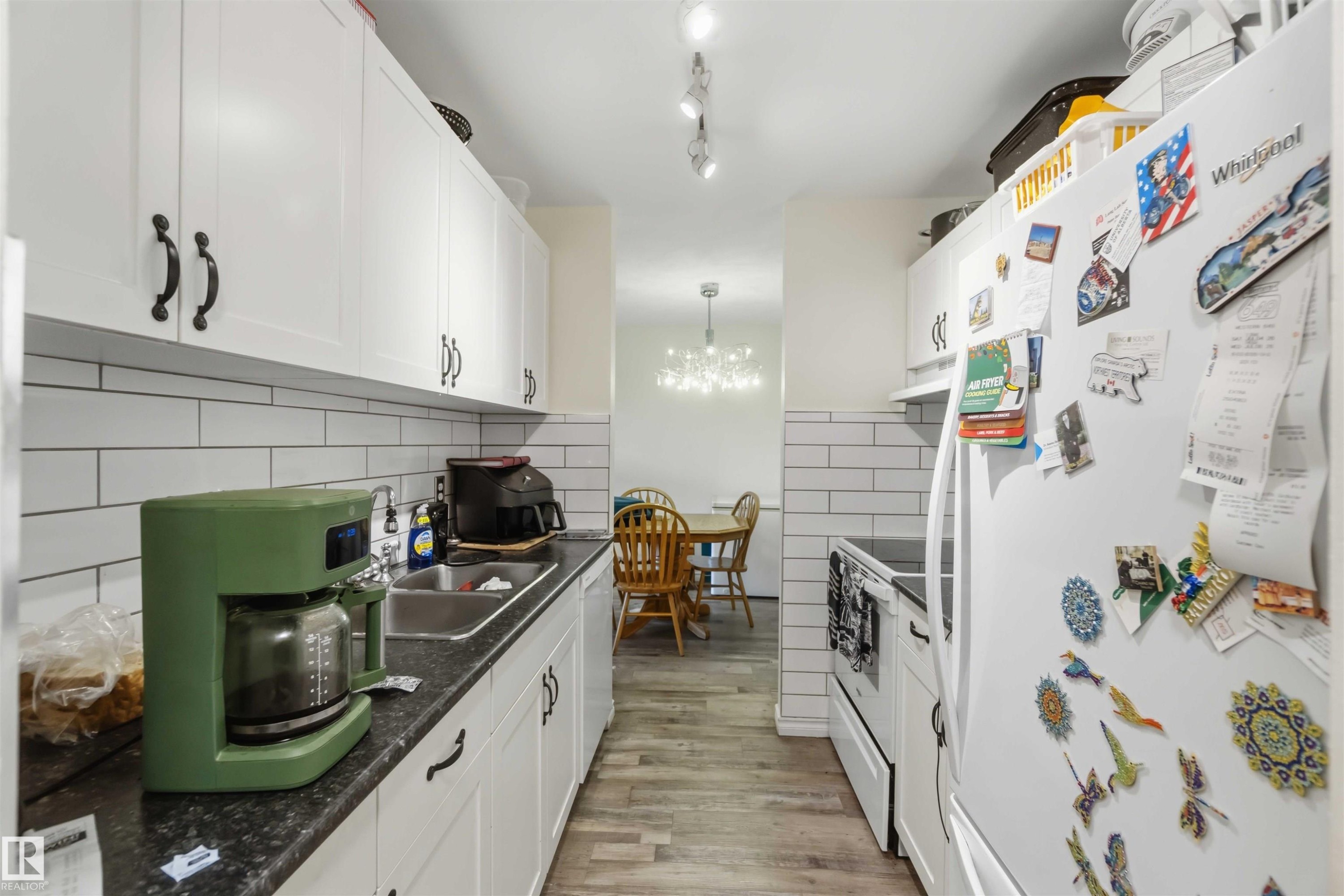 Galley kitchen featuring white cabinetry, black countertops, white subway tile backsplash, and wood-finish flooring - 204 4601 131 Avenue, Edmonton, AB