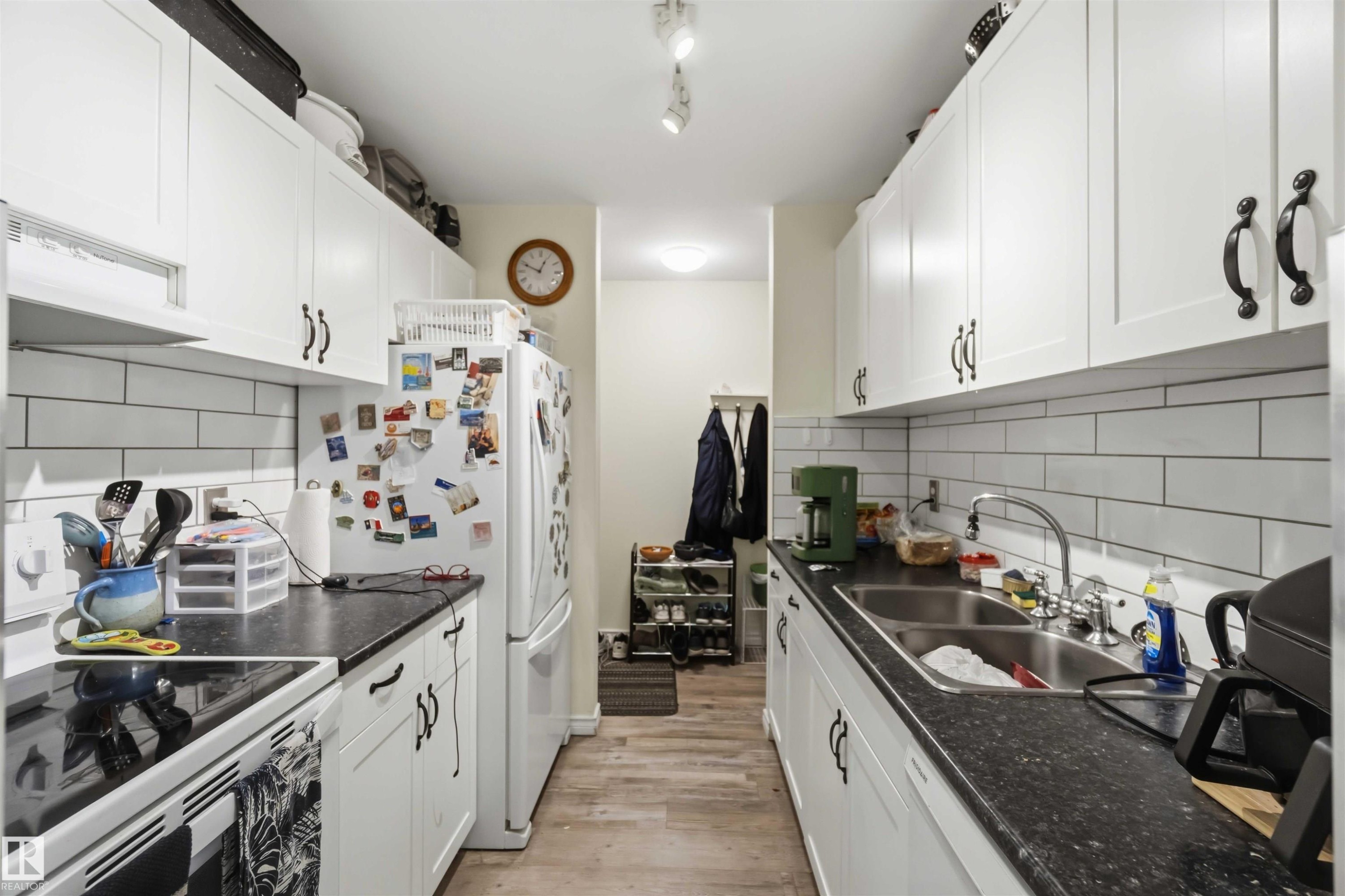 Galley kitchen featuring white cabinetry with black hardware, dark countertops, a subway tile backsplash, and wood-finish flooring - 204 4601 131 Avenue, Edmonton, AB