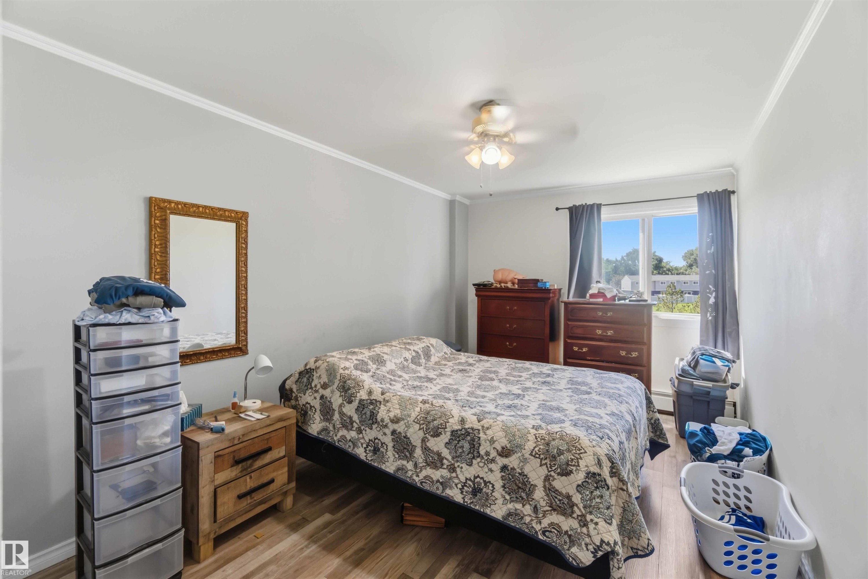 Spacious room featuring light gray walls, white crown molding, wood-finish flooring, a ceiling fan with integrated lighting, and a large window with white trim - 204 4601 131 Avenue, Edmonton, AB