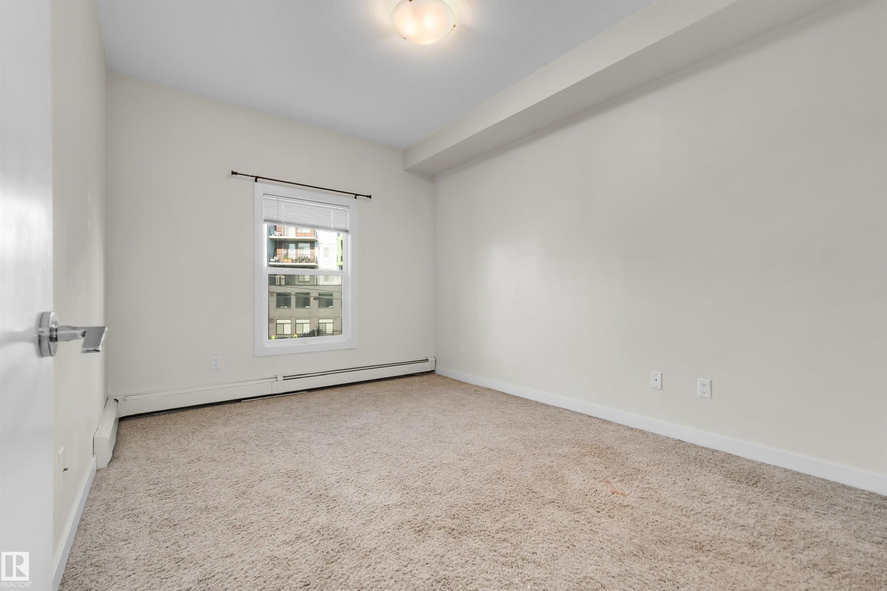 Carpeted room with a single window featuring blinds, a heating unit, and a ceiling-mounted light fixture - #209 10611 117 St Nw, Edmonton, AB - Indoor Photo Showing Other Room