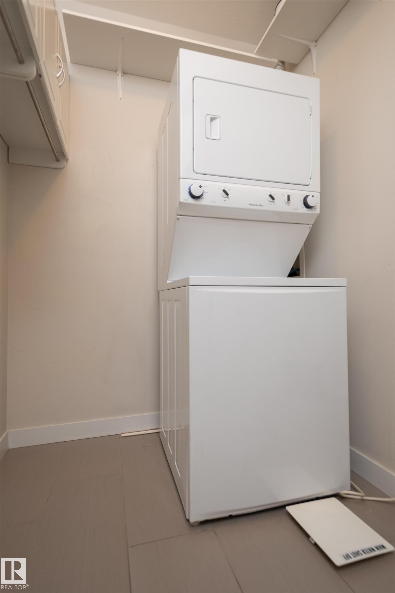 Compact laundry area featuring a white stacked washer and dryer unit, overhead shelving, and neutral wall tones - #209 10611 117 St Nw, Edmonton, AB - Indoor Photo Showing Laundry Room
