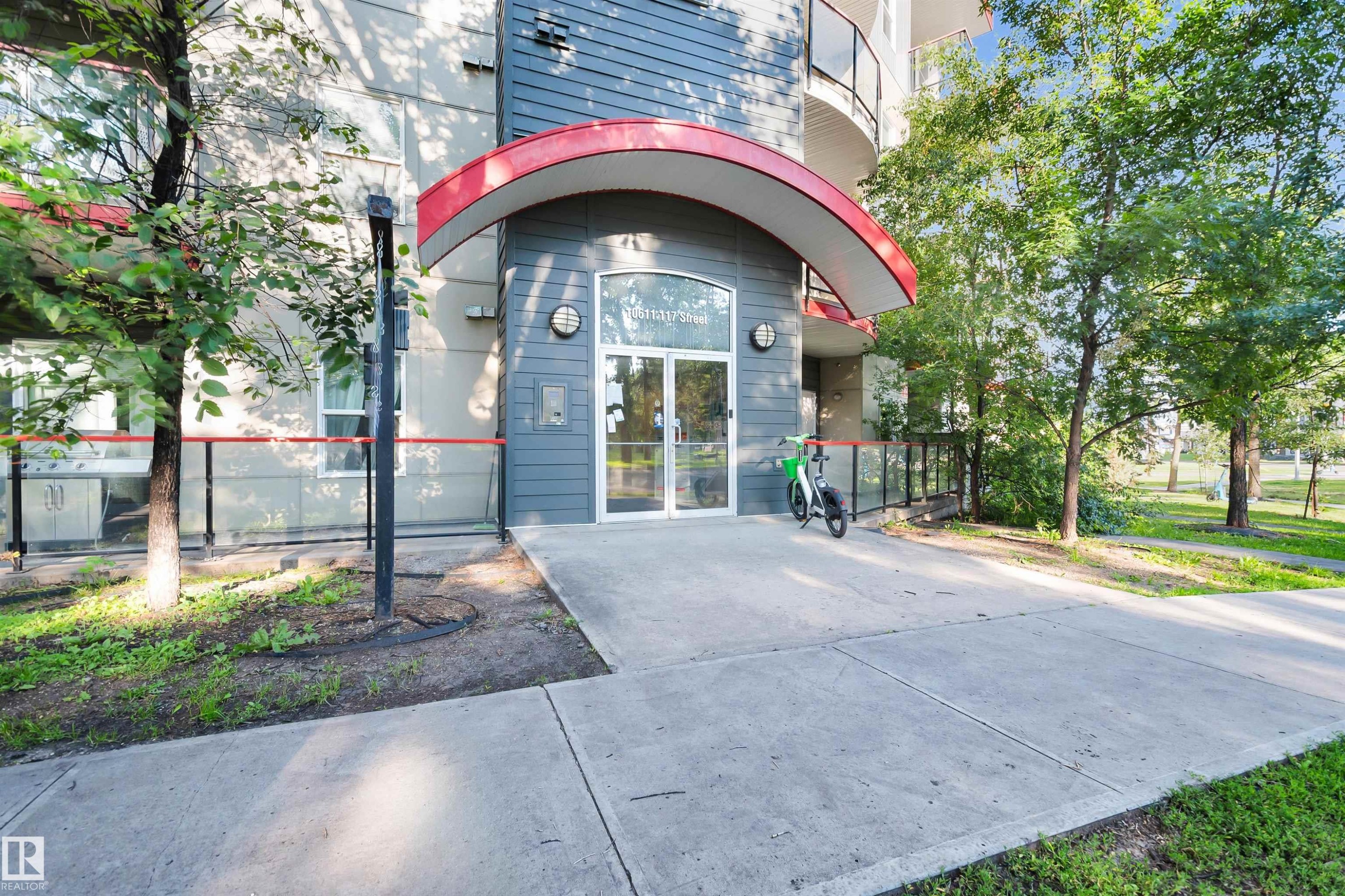 Contemporary building entrance featuring a curved red canopy, grey horizontal siding, and a glass-paneled entry door - #209 10611 117 St Nw, Edmonton, AB - Outdoor