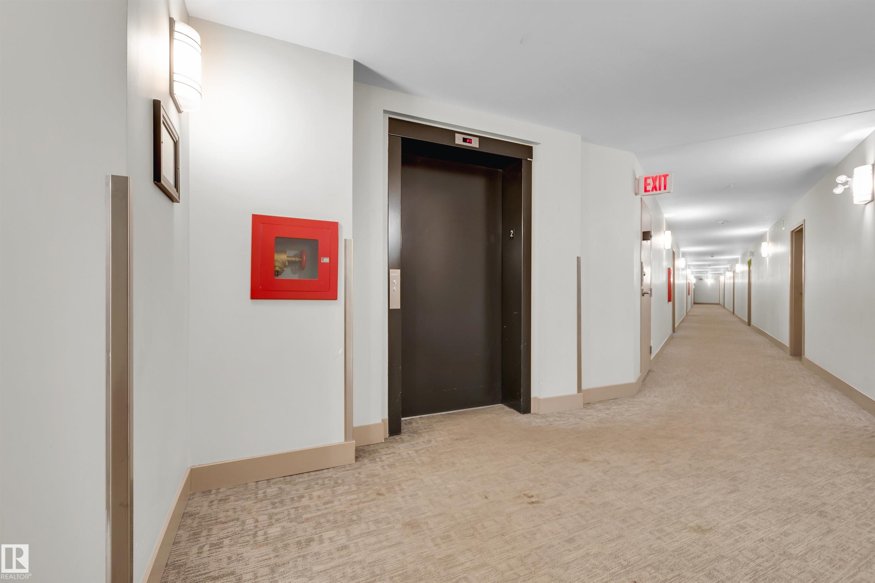 Building corridor featuring neutral-toned carpet flooring, wall-mounted light fixtures, white walls, and a dark elevator door - #209 10611 117 St Nw, Edmonton, AB - Indoor Photo Showing Other Room