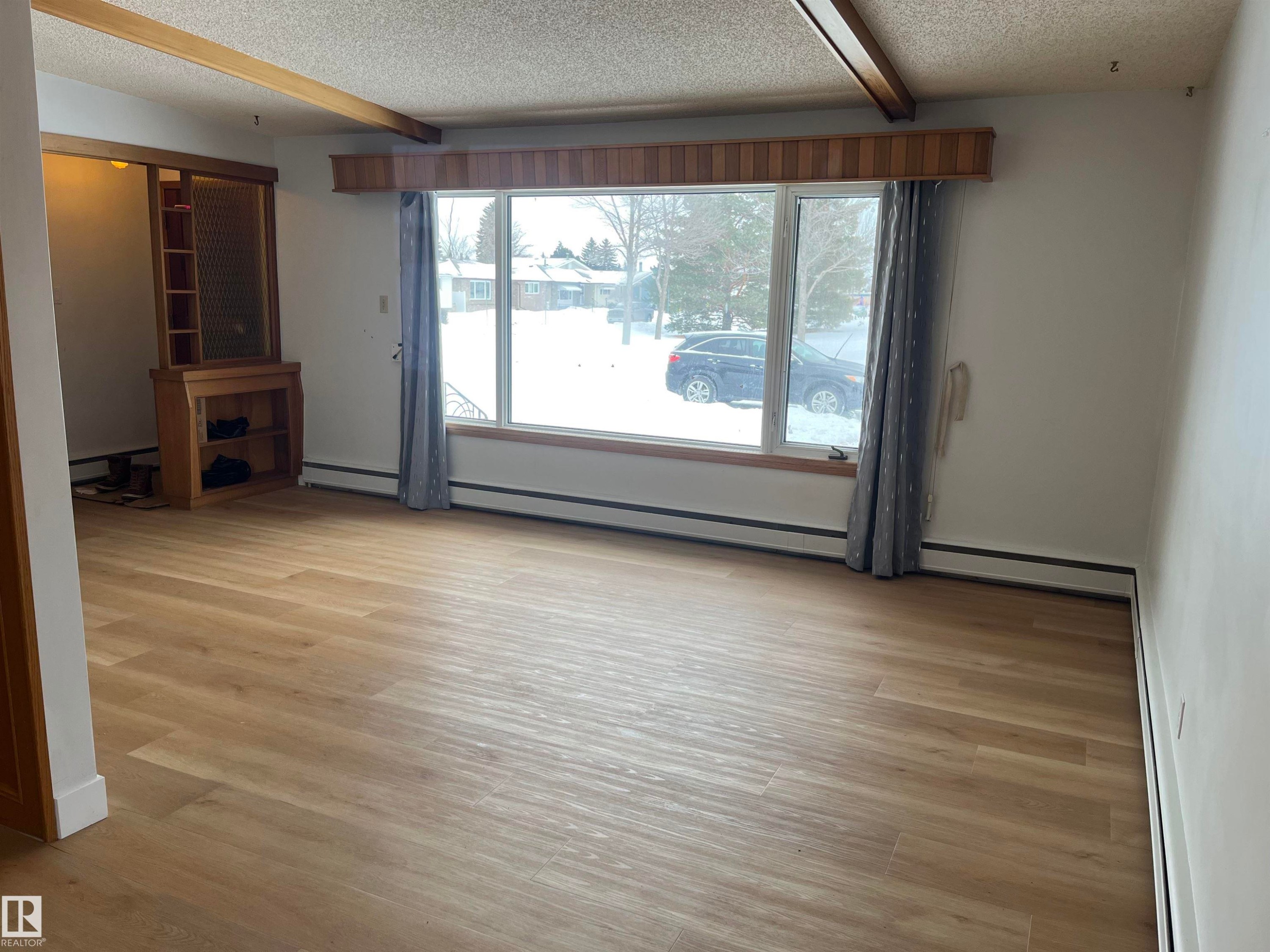 Spacious living area featuring light wood-finish flooring, a large picture window with a wood valance, and a built-in wood shelving unit - 8736 72 Street Nw, Edmonton, AB - Indoor Photo Showing Other Room