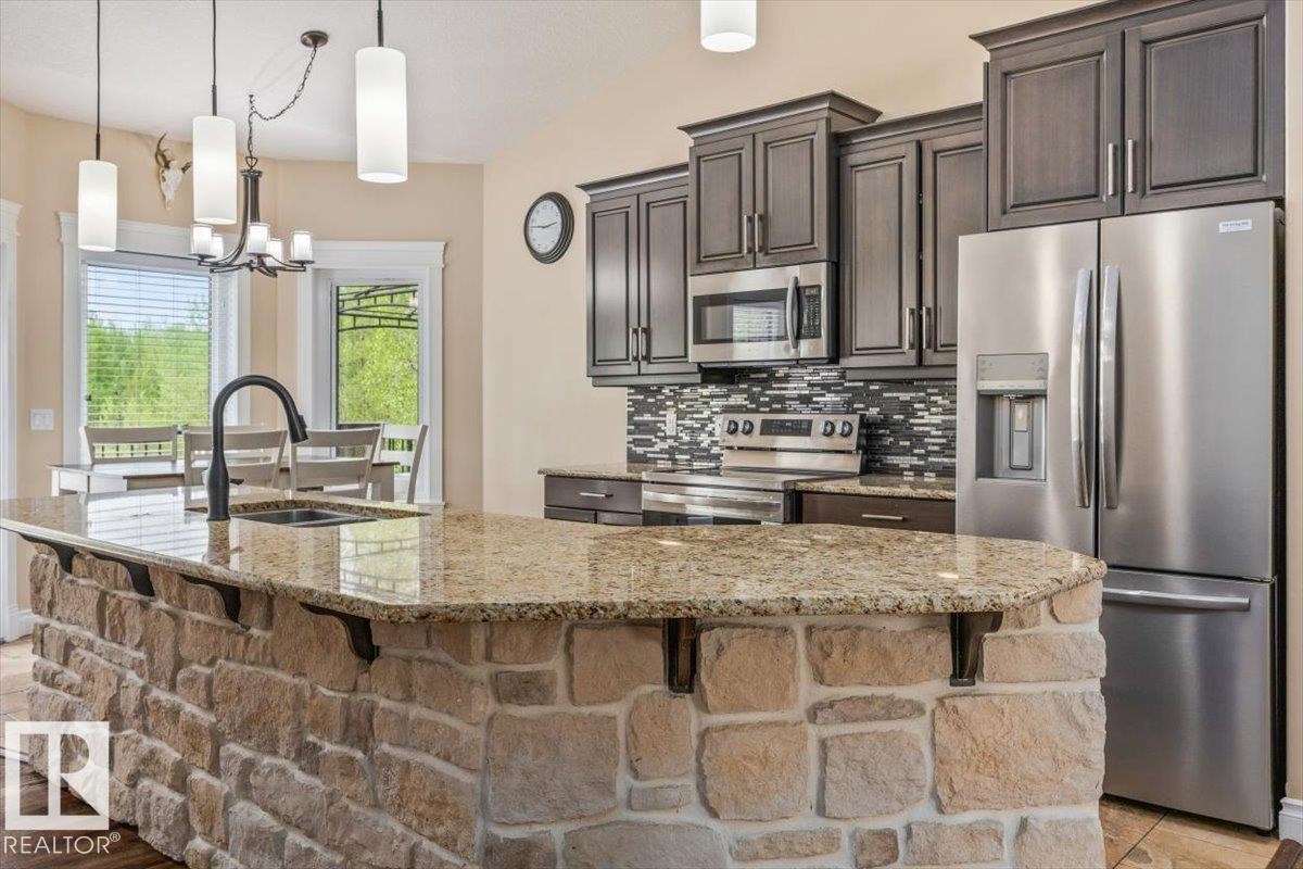 Kitchen featuring a stone-faced island with a granite-style countertop, stainless steel appliances, dark wood-finish cabinetry, a mosaic tile backsplash, and pendant lighting - #6 2406 Twp Road 540, Rural Lac Ste. Anne County, AB - Indoor Photo Showing Kitchen With Double Sink With Upgraded Kitchen
