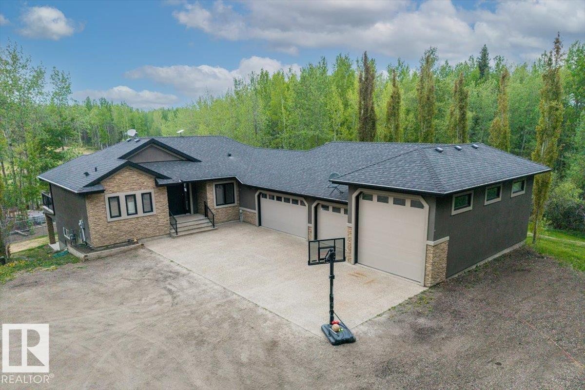 Modern exterior featuring stone accents, dark siding, multiple garage doors, a concrete driveway, and surrounding mature trees - #6 2406 Twp Road 540, Rural Lac Ste. Anne County, AB - Outdoor