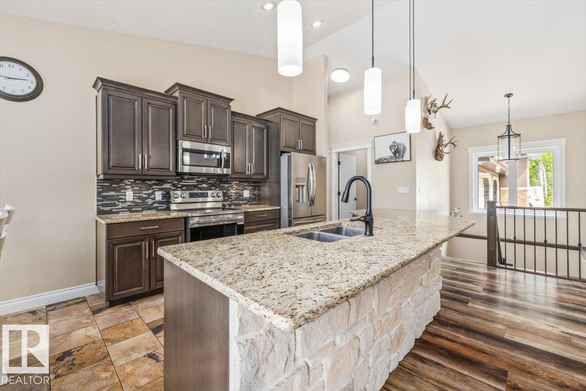 Kitchen with dark wood cabinetry, stainless steel appliances, a mosaic tile backsplash, and a stone-faced island featuring a double sink and light-toned countertop - #6 2406 Twp Road 540, Rural Lac Ste. Anne County, AB - Indoor Photo Showing Kitchen With Double Sink With Upgraded Kitchen