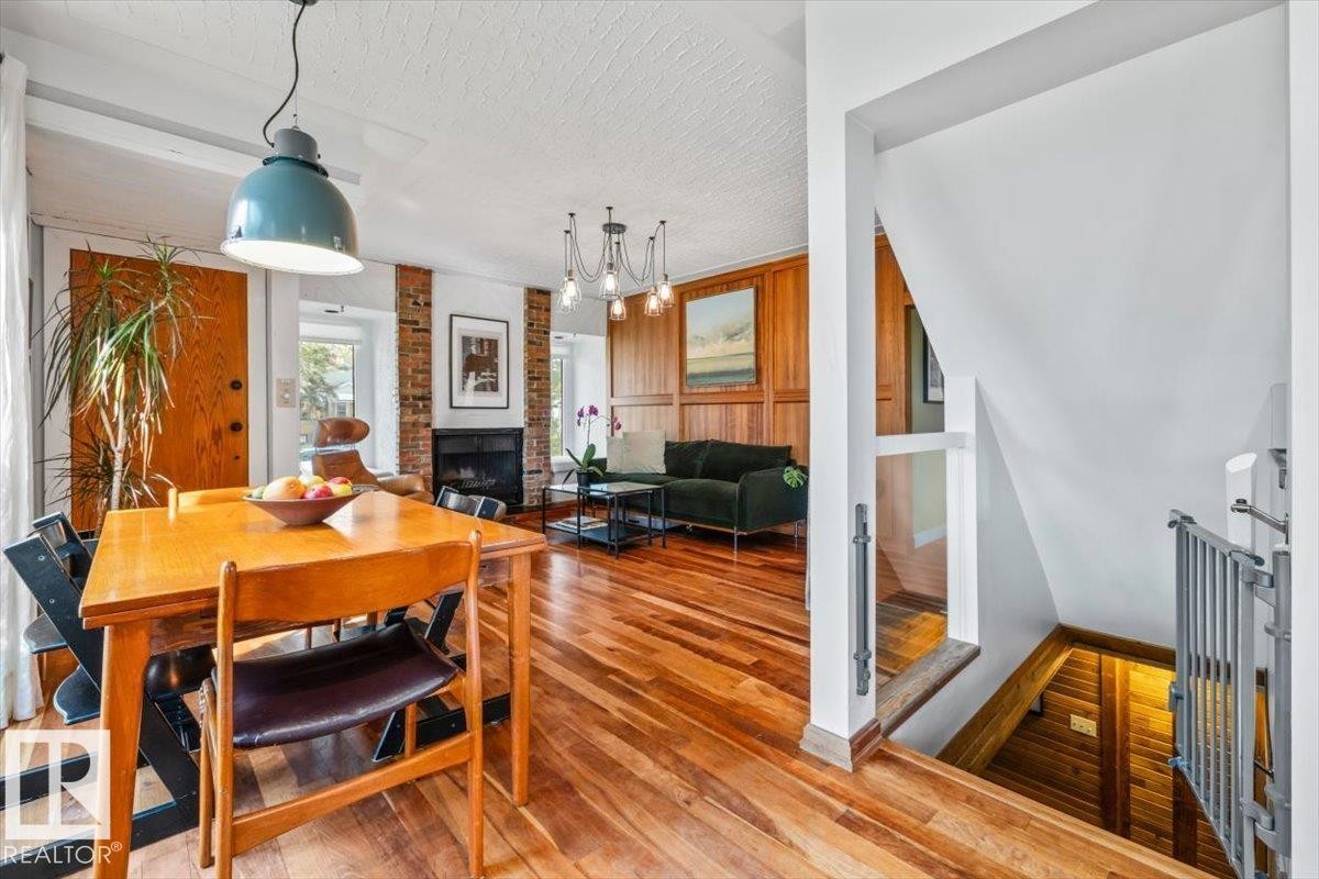 Open-concept living space with wood-finish flooring, a brick accent wall, wood paneling, a built-in fireplace, and a staircase with metal railings - 11603 70 Street Nw, Edmonton, AB