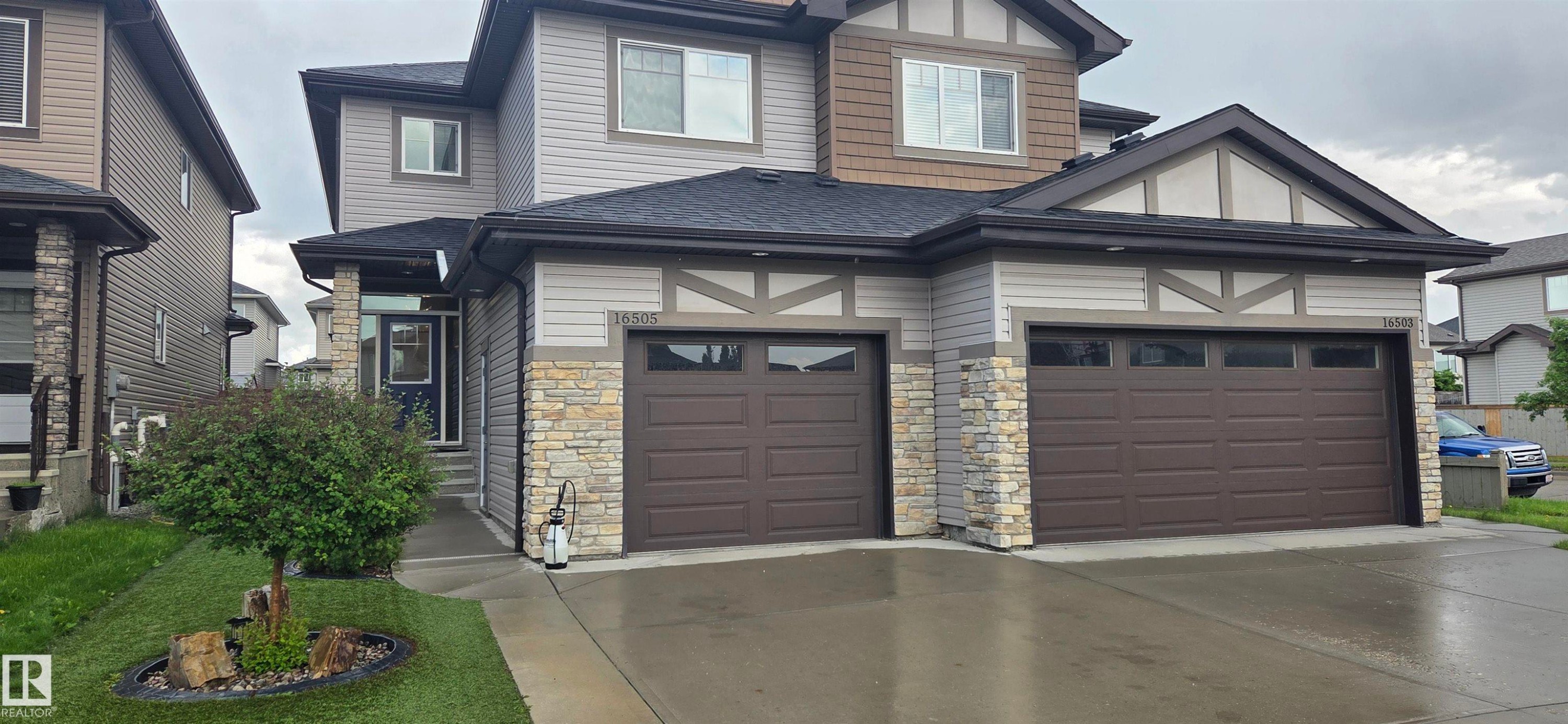 Contemporary duplex featuring light-toned siding, stone veneer accents, dark shingle roofing, dark brown garage doors with upper window panels, and a concrete driveway - 16505 133 Street, Edmonton, AB