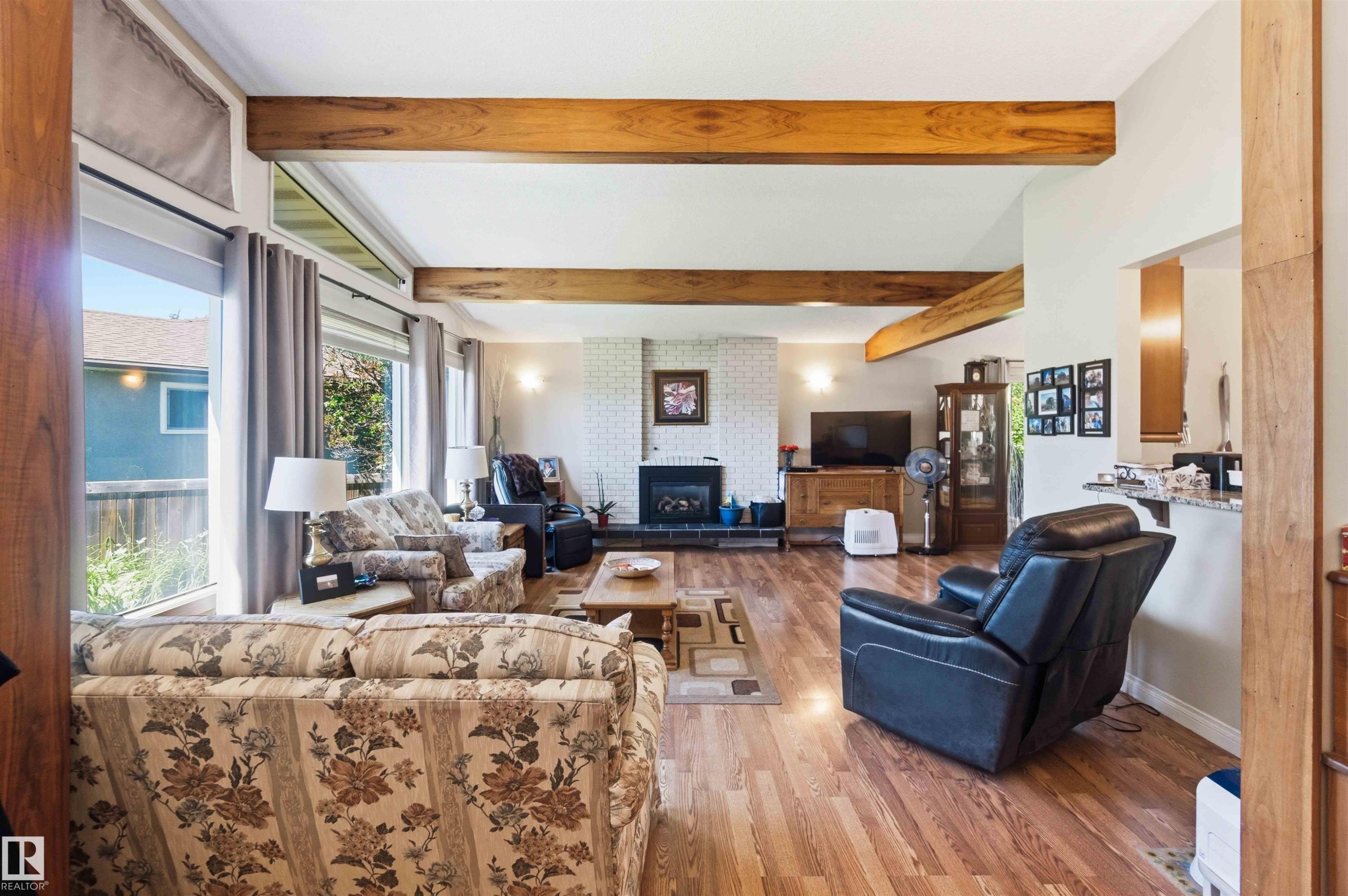 Living area featuring wood-finish flooring, exposed ceiling beams, a white brick fireplace, and large picture windows - 13121 122 Street, Edmonton, AB