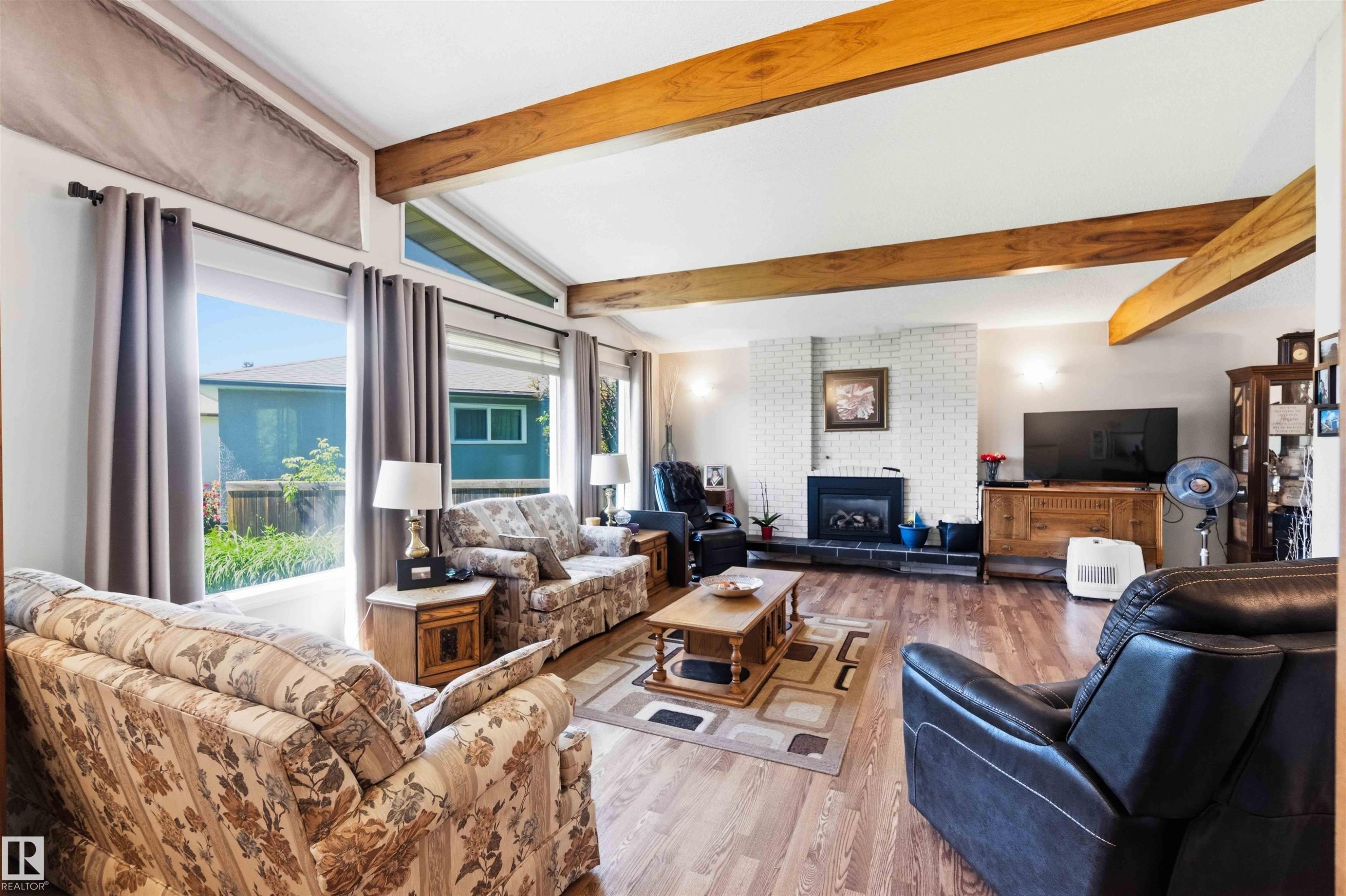 Spacious living area featuring exposed wood beams, wood-finish flooring, and a white brick fireplace with a black hearth - 13121 122 Street, Edmonton, AB