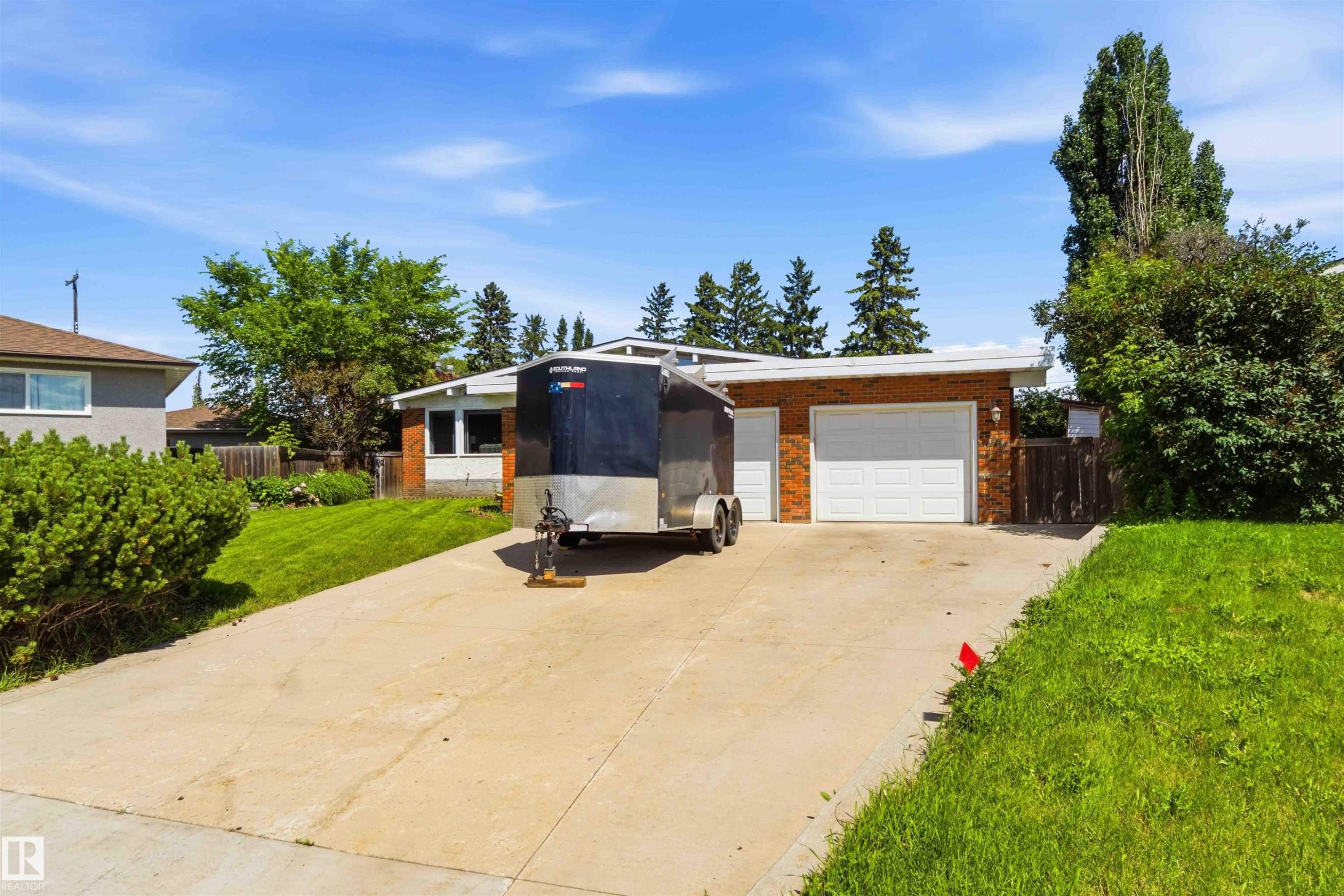 Brick exterior with a white garage door and a concrete driveway, complemented by mature landscaping and a grassy front yard - 13121 122 Street, Edmonton, AB