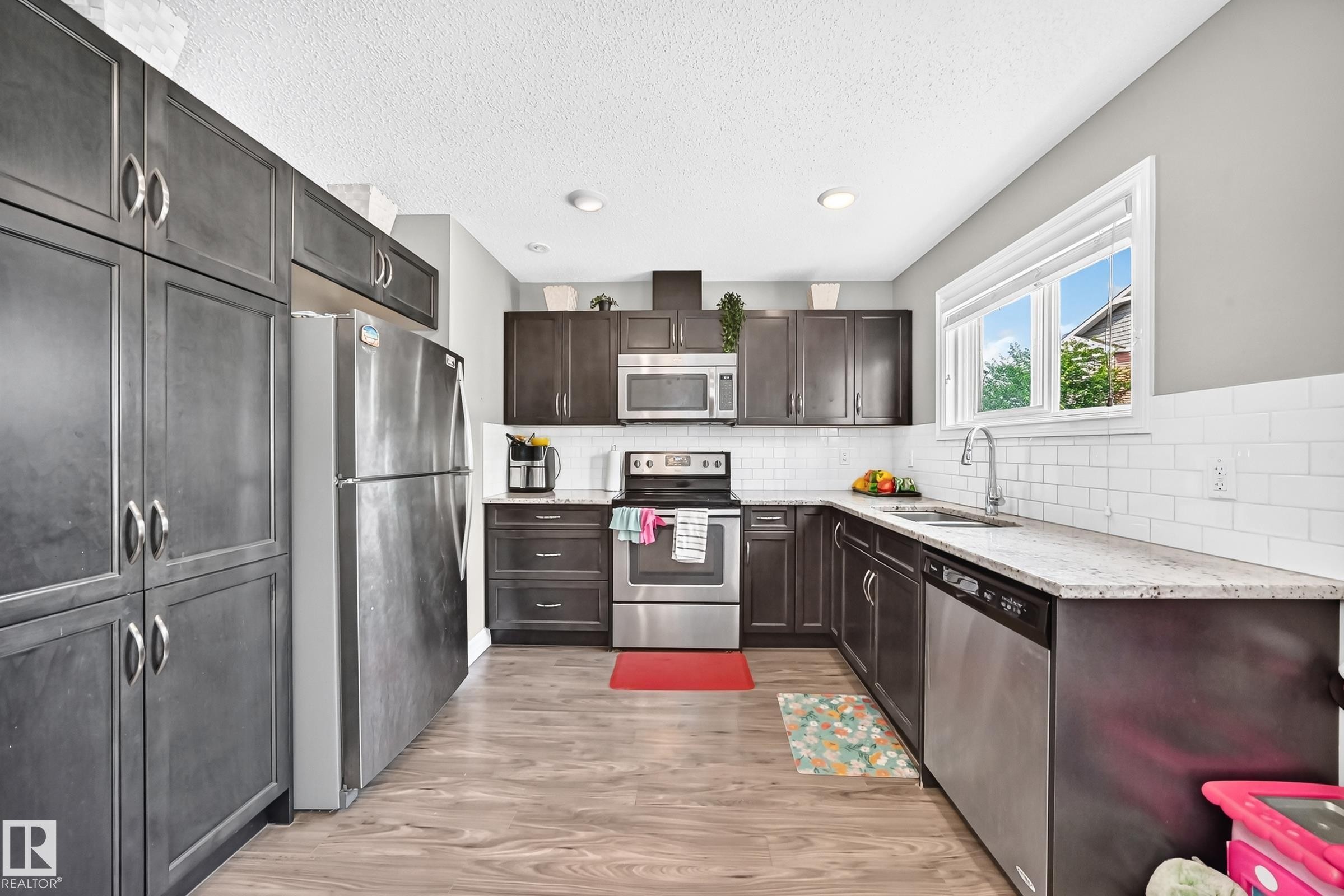 Modern kitchen featuring dark cabinetry, stainless steel appliances, light-toned countertops, a white subway tile backsplash, and wood-finish flooring - 415 Clareview Rd Nw Nw, Edmonton, AB - Indoor Photo Showing Kitchen With Upgraded Kitchen