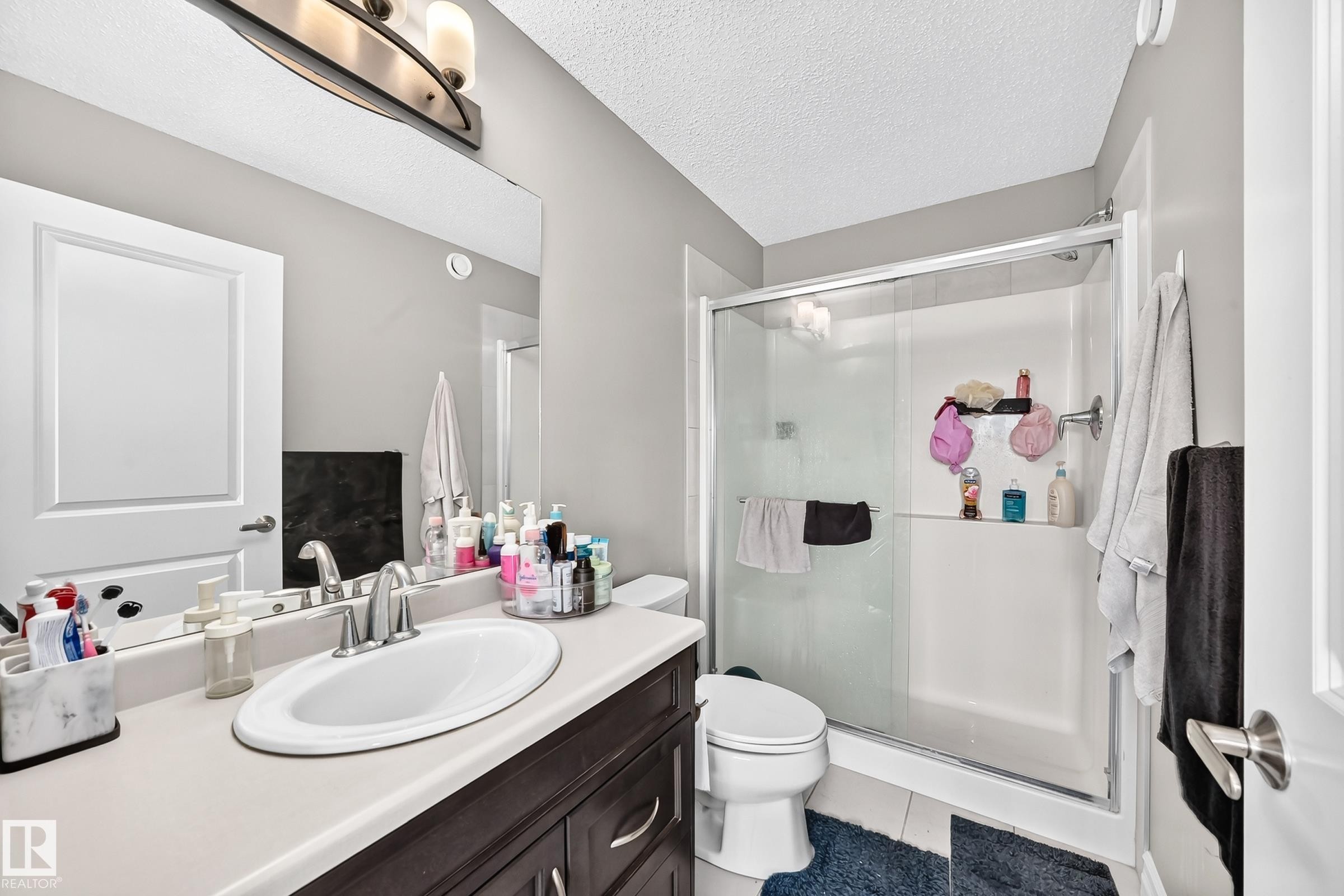 Bathroom featuring a vanity with an integrated sink, a large wall-mounted mirror, a modern three-light fixture, a toilet, and a glass-enclosed shower with a built-in shelf - 415 Clareview Rd Nw Nw, Edmonton, AB - Indoor Photo Showing Bathroom