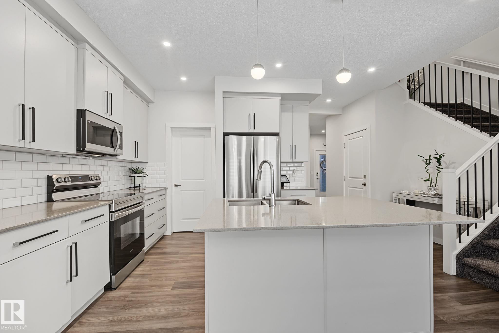 Contemporary kitchen featuring a large island with an undermount sink, white cabinetry with black hardware, stainless steel appliances, and wood-finish flooring - 432 Roberts Crescent, Leduc, AB