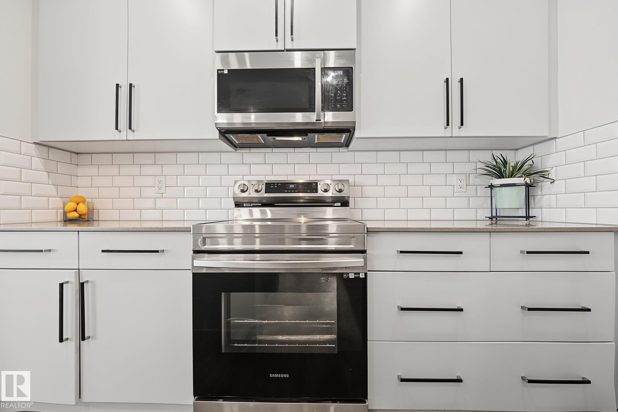 Contemporary kitchen featuring white cabinetry with black bar pulls, stainless steel appliances, a white subway tile backsplash, and light-toned countertops - 432 Roberts Crescent, Leduc, AB