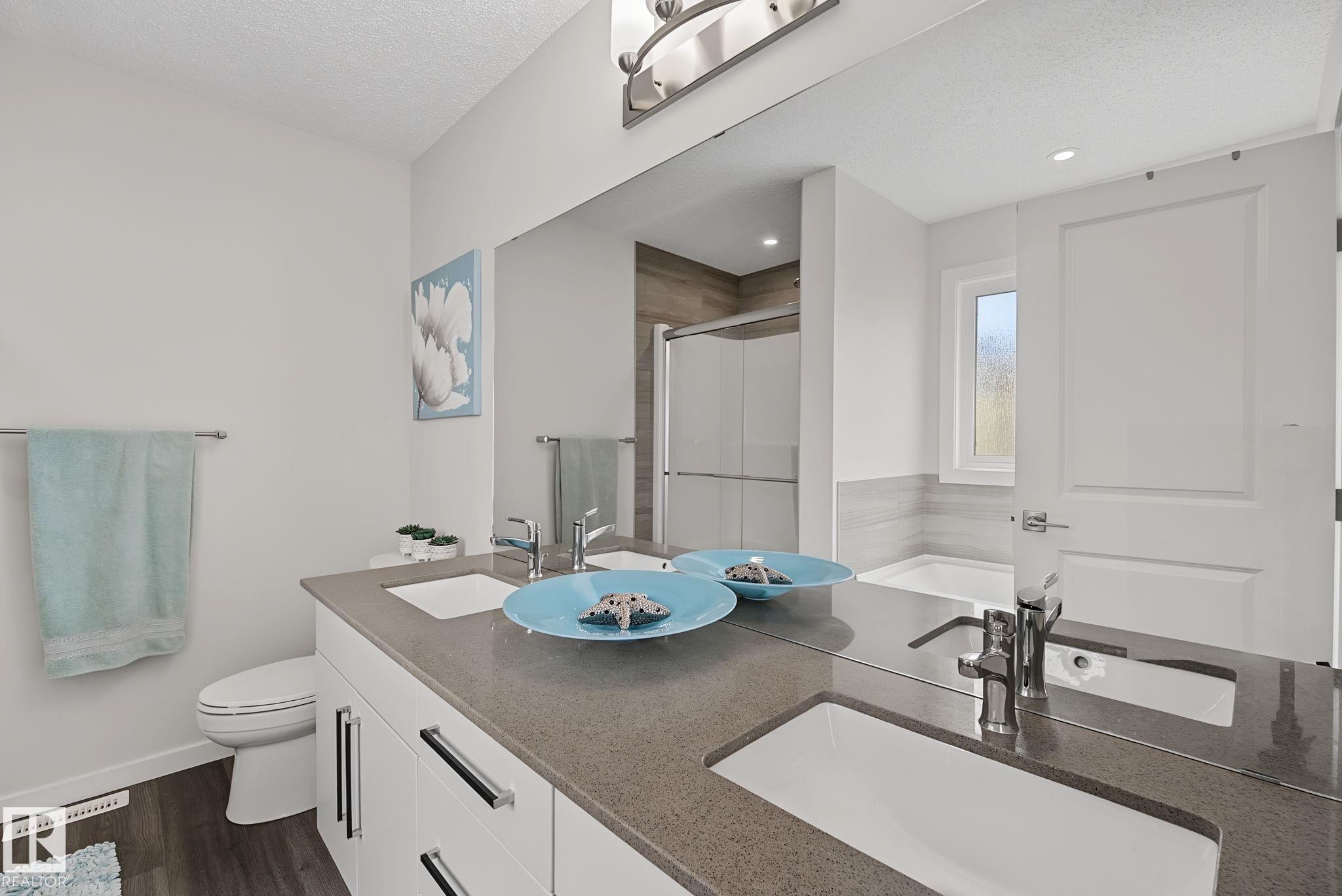 Bathroom featuring a dual vanity with undermount sinks and chrome fixtures, a large wall-mounted mirror, wood-finish flooring, and a window above the soaking tub - 432 Roberts Crescent, Leduc, AB