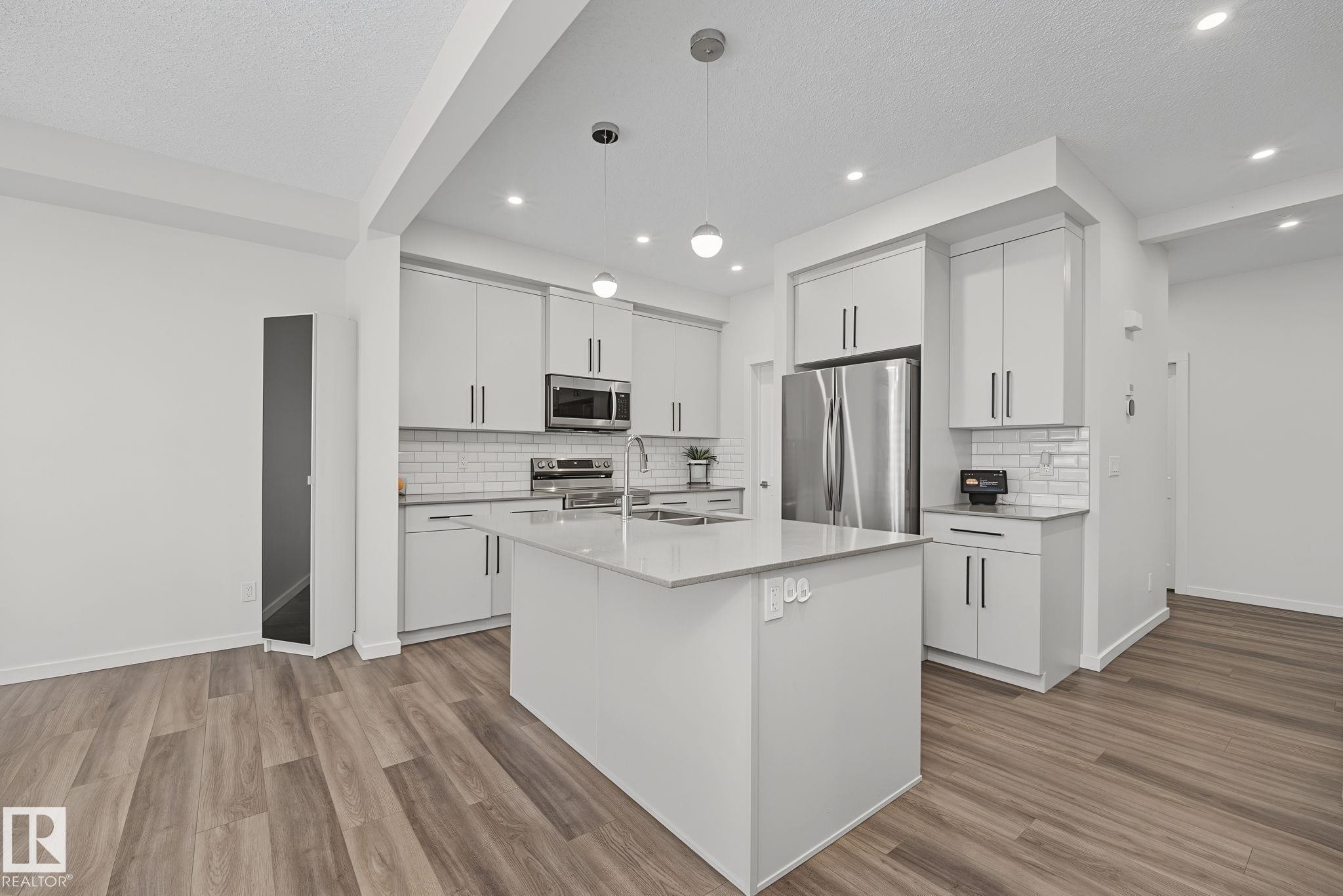 Modern kitchen featuring light gray cabinetry, stainless steel appliances, a center island with a sink, white subway tile backsplash, and wood-finish flooring - 432 Roberts Crescent, Leduc, AB