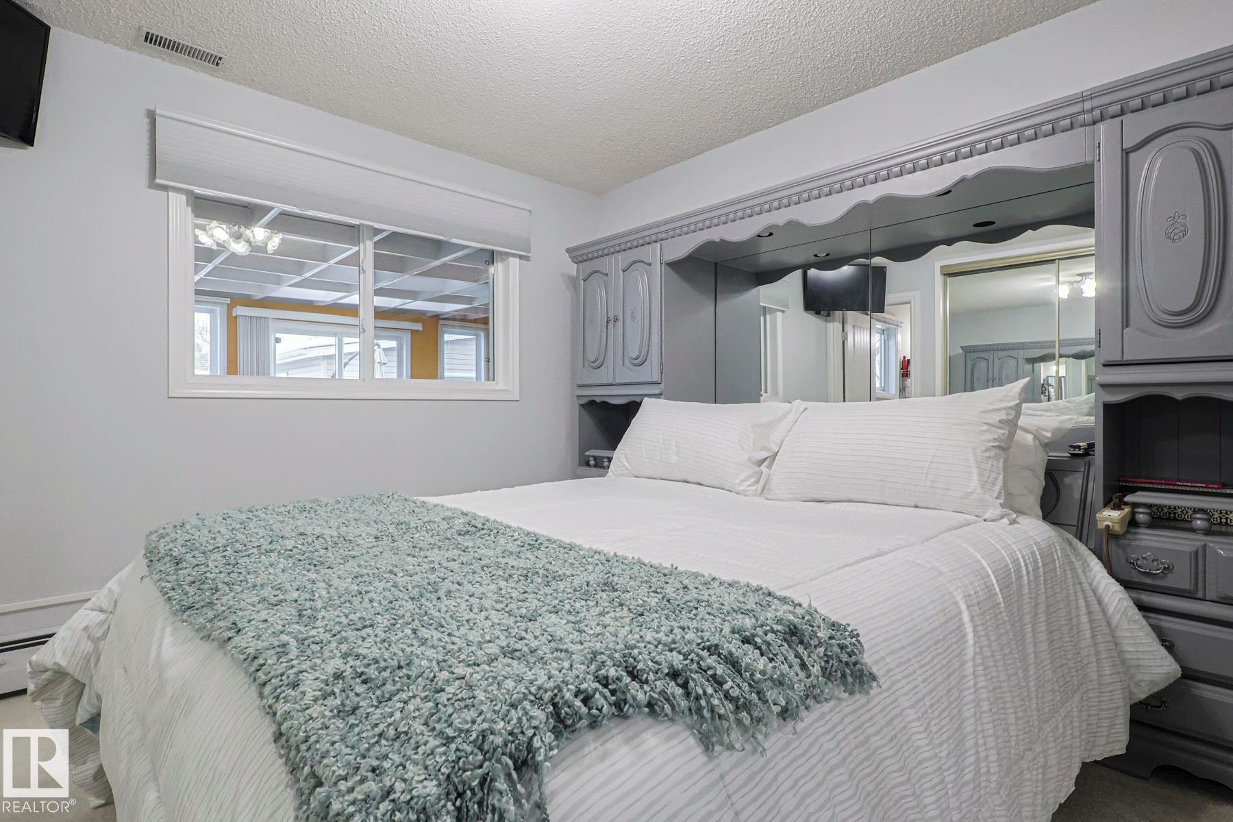Bedroom featuring a multi-pane window with a roller shade, light gray walls, built-in mirrored cabinetry, and textured ceiling - 13304 70 Street, Edmonton, AB