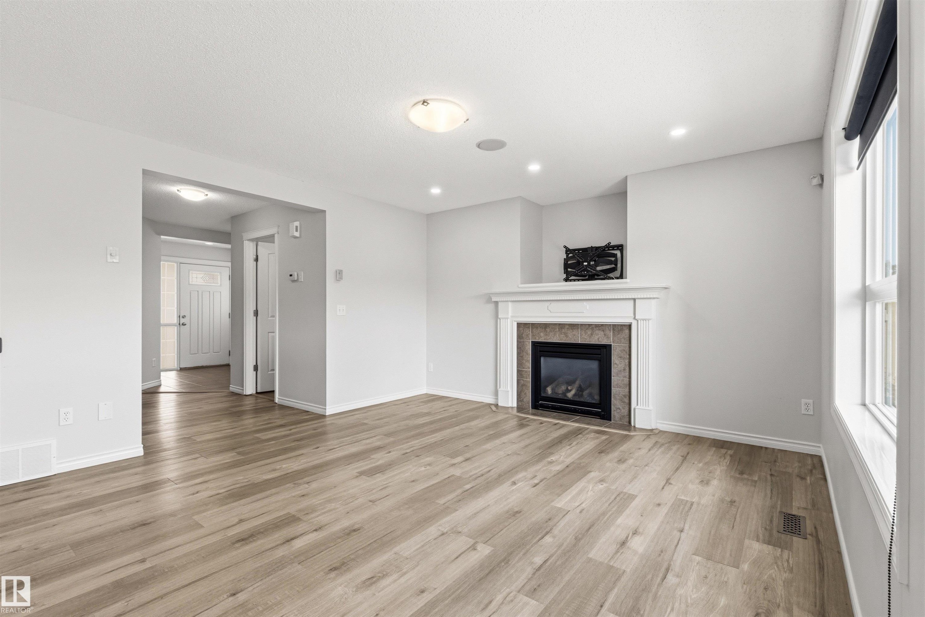 Spacious living area featuring wood-finish flooring, a contemporary fireplace with a white mantel and tiled surround, multiple recessed lights, a large window, and a ceiling-mounted light fixture - 3214 22A Avenue Nw, Edmonton, AB - Indoor Photo Showing Living Room With Fireplace