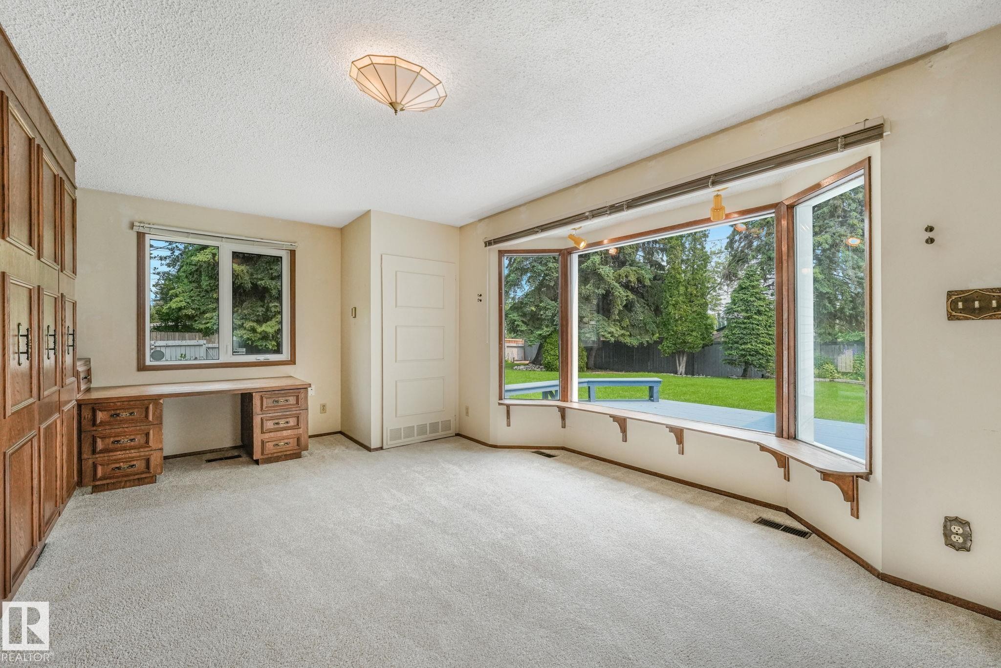 Spacious room featuring a large bay window overlooking a green yard, built-in desk with drawers, extensive wood-paneled cabinetry, and light-colored carpeting - 13320 79 Avenue Nw, Edmonton, AB - Indoor