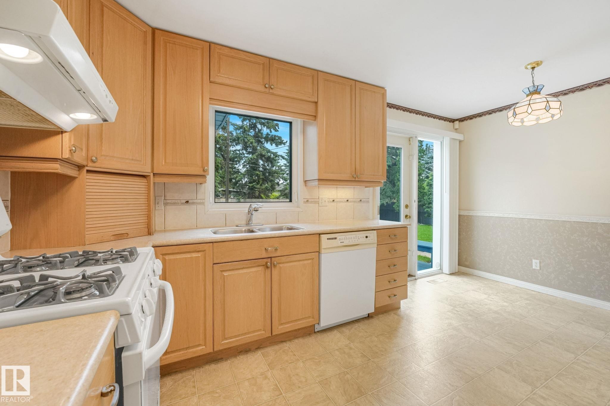 Kitchen featuring natural wood-finish cabinetry, a window overlooking greenery, integrated double basin sink, and a white dishwasher - 13320 79 Avenue Nw, Edmonton, AB - Indoor Photo Showing Kitchen With Double Sink