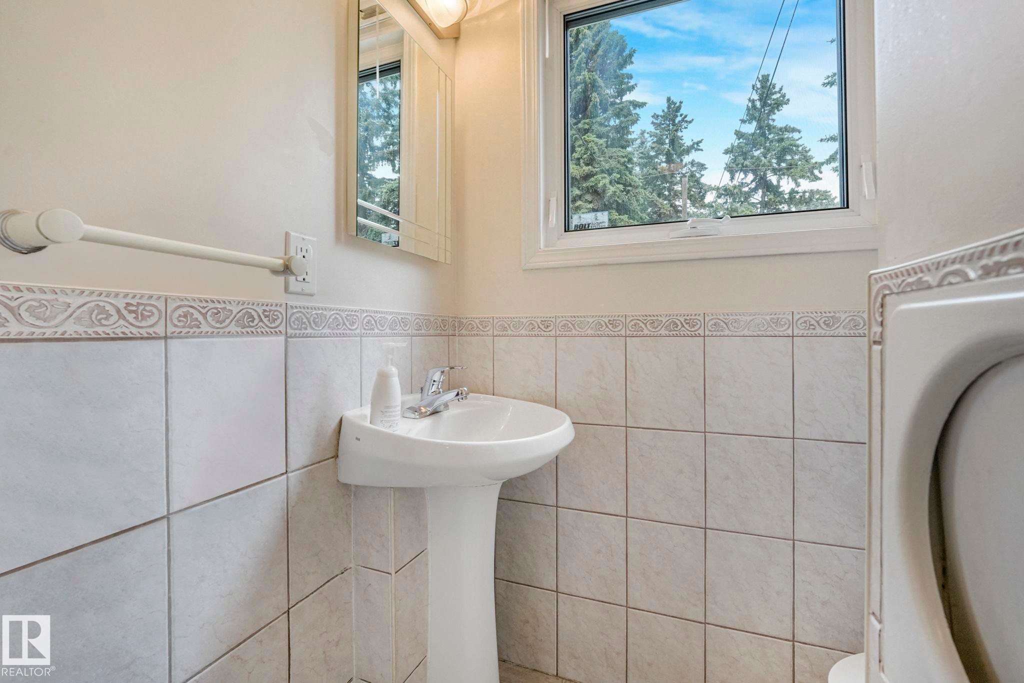 Bathroom featuring light-toned tile wainscoting with decorative border, pedestal sink with chrome faucet, and a window framing mature trees - 13320 79 Avenue Nw, Edmonton, AB - Indoor Photo Showing Bathroom