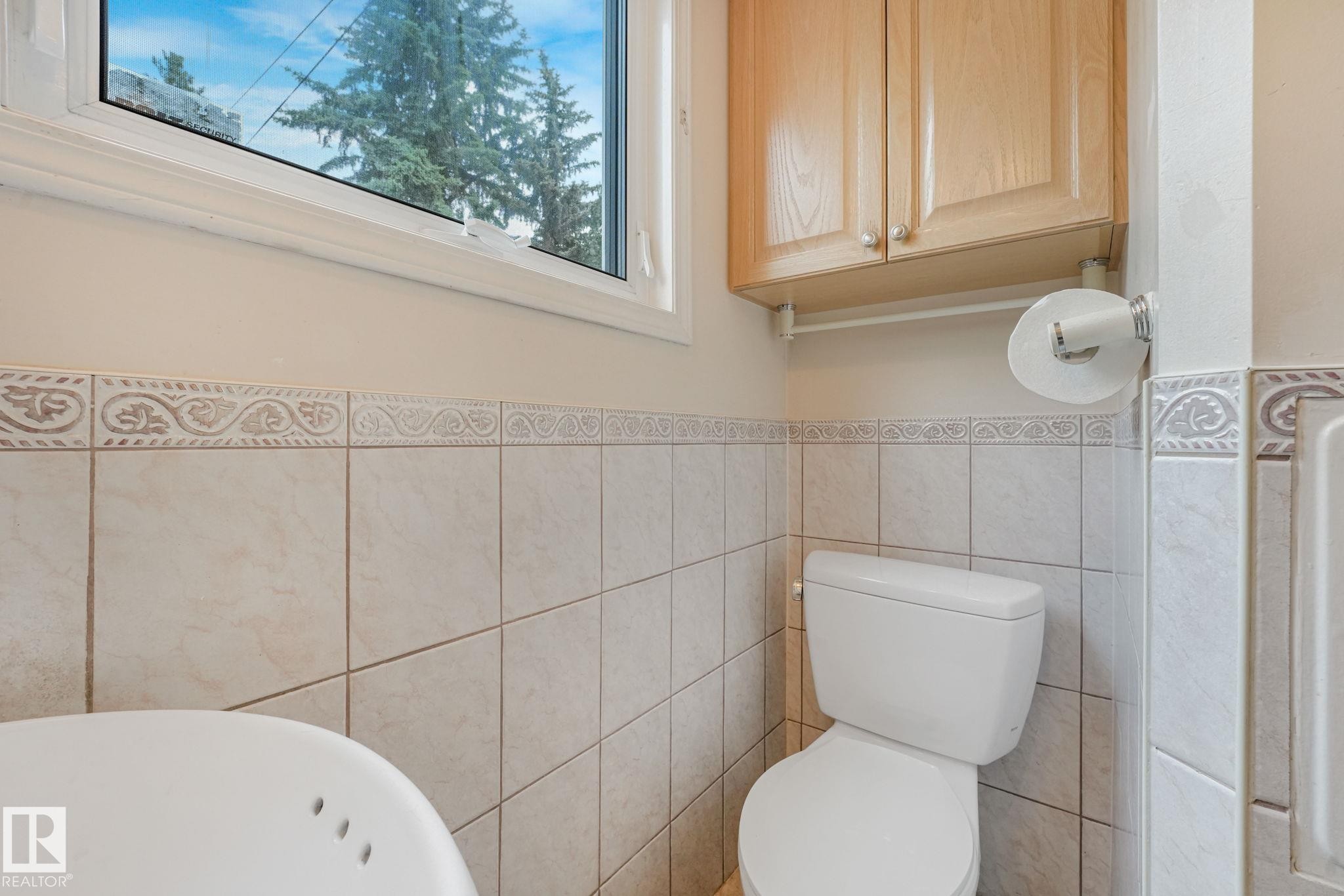 Bathroom featuring light-toned square wall tiles, decorative accent border tiles, and a window with white trim - 13320 79 Avenue Nw, Edmonton, AB - Indoor Photo Showing Bathroom