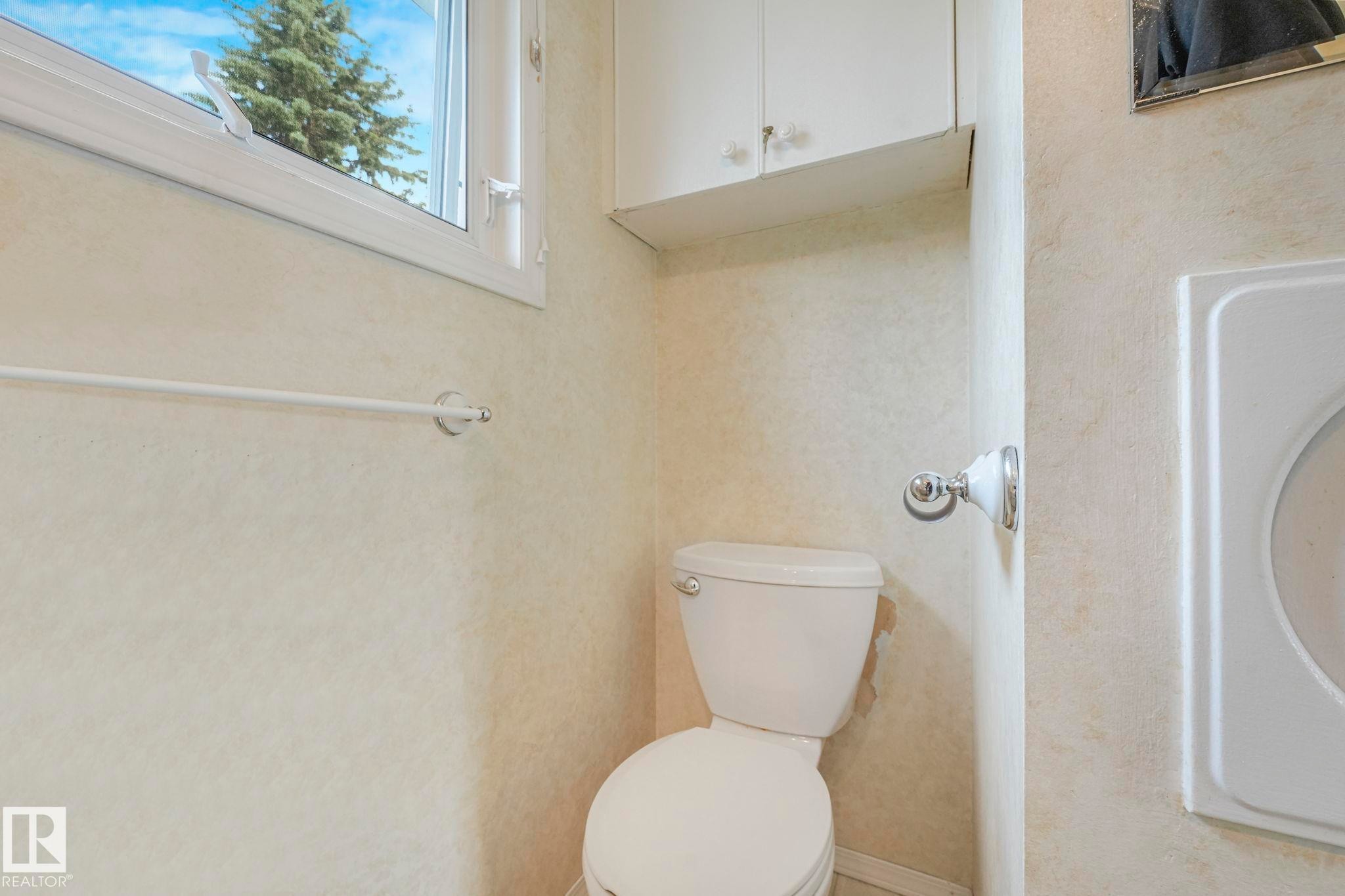 White toilet with overhead cabinetry, textured wall finish, and a window with a tree background - 13320 79 Avenue Nw, Edmonton, AB - Indoor Photo Showing Bathroom