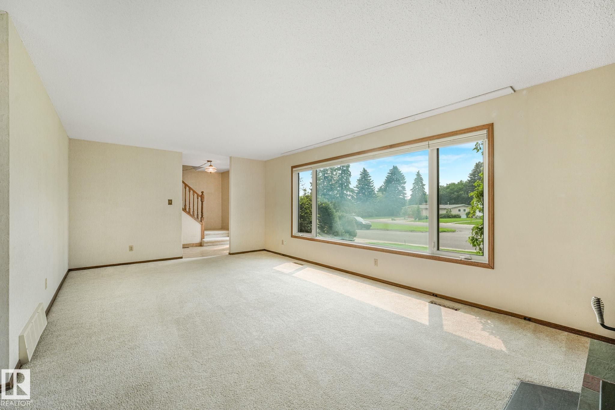 Carpeted living area featuring a large picture window with wood-finish trim, light-colored walls, and a visible staircase with wooden railings and risers - 13320 79 Avenue Nw, Edmonton, AB - Indoor Photo Showing Other Room