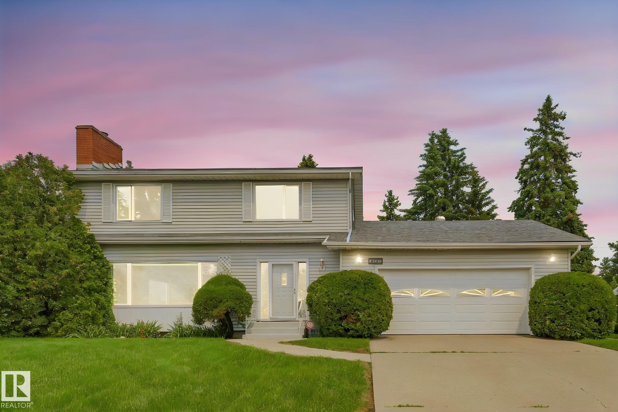 Two-story residence featuring light-colored siding, a prominent brick chimney, and a two-car garage with overhead lighting - 13320 79 Avenue Nw, Edmonton, AB - Outdoor