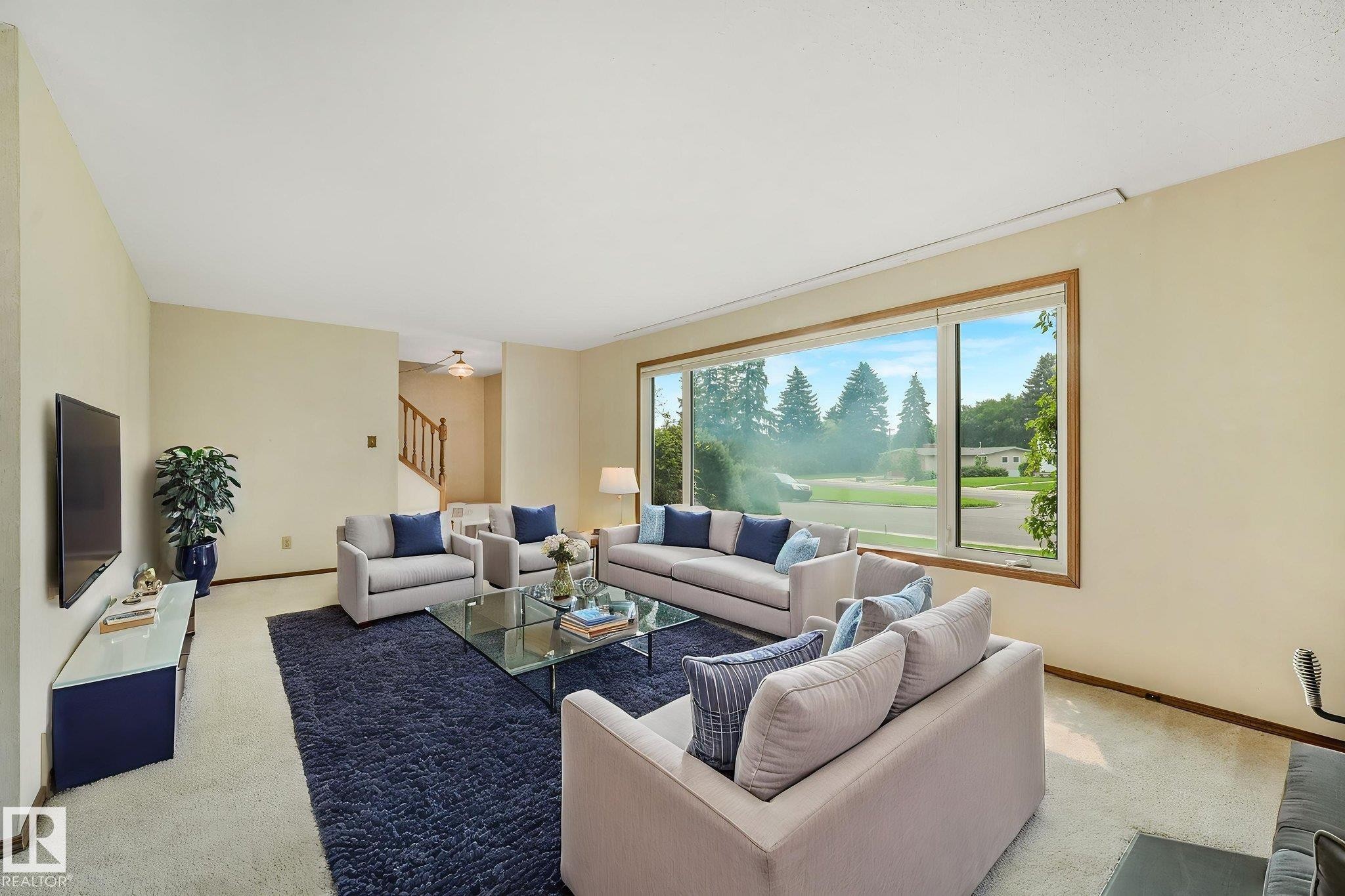 Expansive living area featuring a large picture window with wood trim, light-toned walls, and a staircase with wood railings visible in the background - 13320 79 Avenue Nw, Edmonton, AB - Indoor Photo Showing Living Room