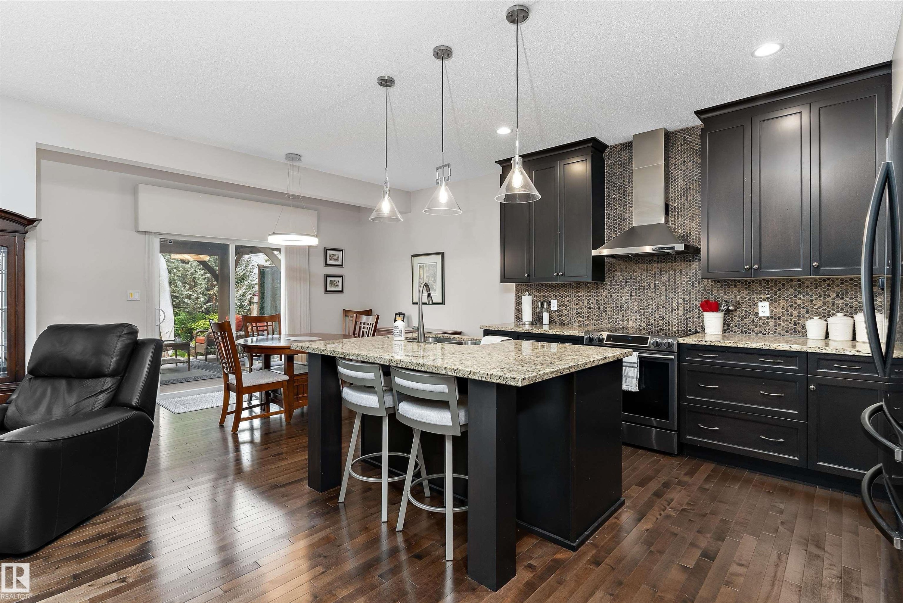 Open concept kitchen featuring dark wood cabinetry, a granite-topped island with seating, a mosaic tile backsplash, stainless steel range hood, and wood-finish flooring - 1097 Armitage Crescent, Edmonton, AB