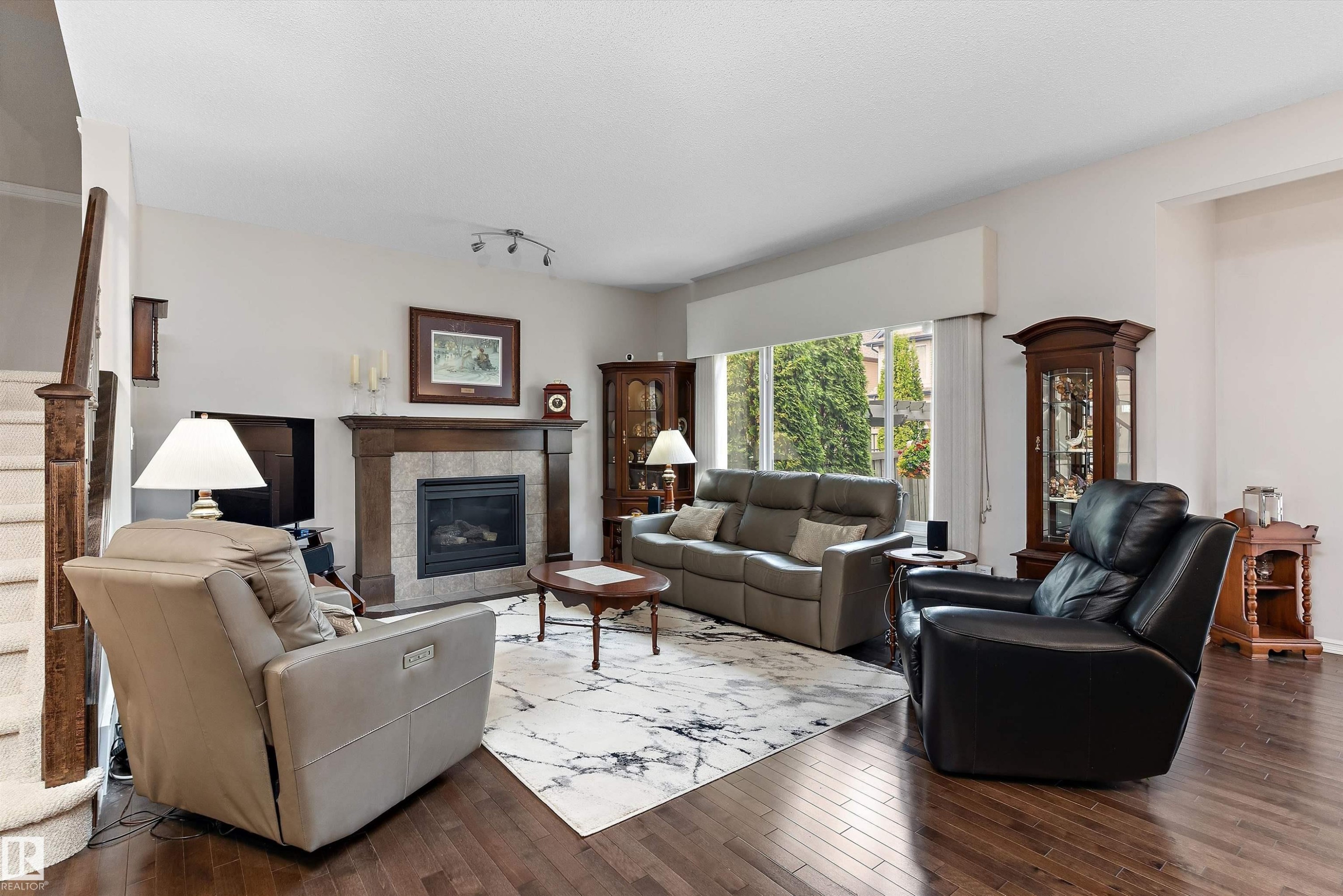 Living area featuring dark wood-finish flooring, a fireplace with a stone surround and wood mantel, a large window with exterior greenery, and a staircase with wood newel posts and carpeted treads - 1097 Armitage Crescent, Edmonton, AB