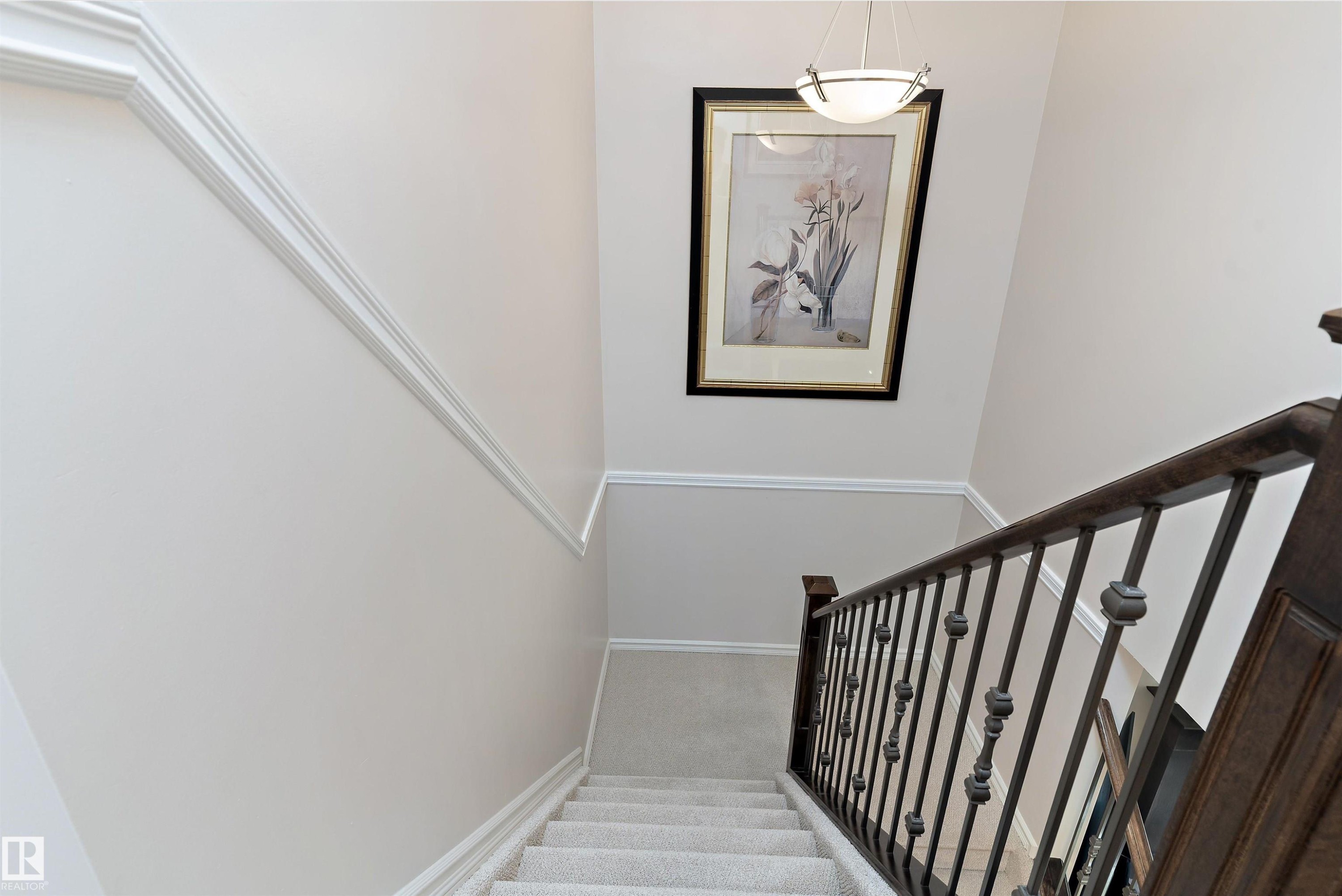Carpeted staircase featuring a dark wood handrail with wrought iron balusters, white trim, and a high ceiling with a pendant light fixture - 1097 Armitage Crescent, Edmonton, AB