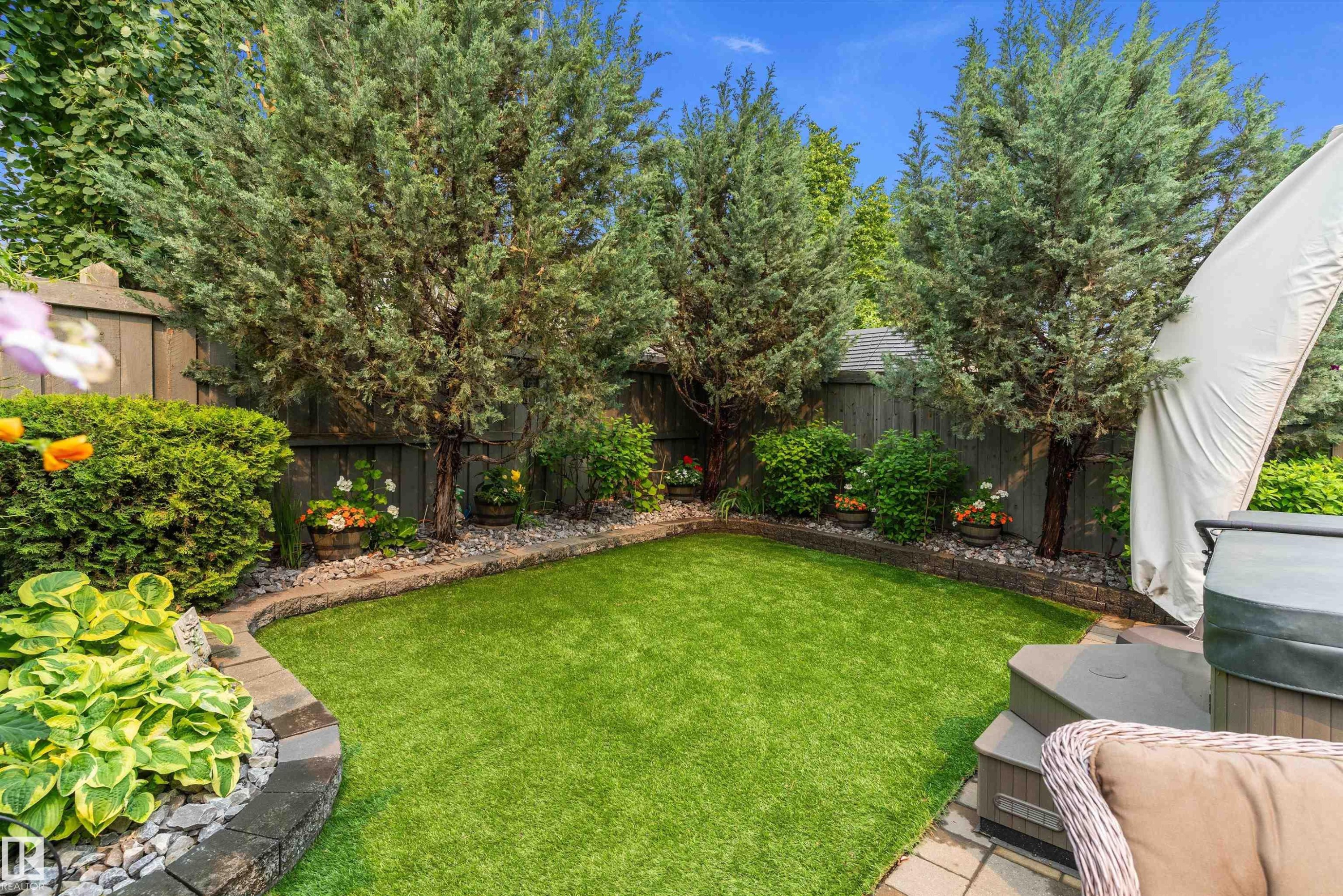 Backyard featuring artificial turf, a stone-bordered garden bed, mature evergreen trees, and a privacy fence - 1097 Armitage Crescent, Edmonton, AB