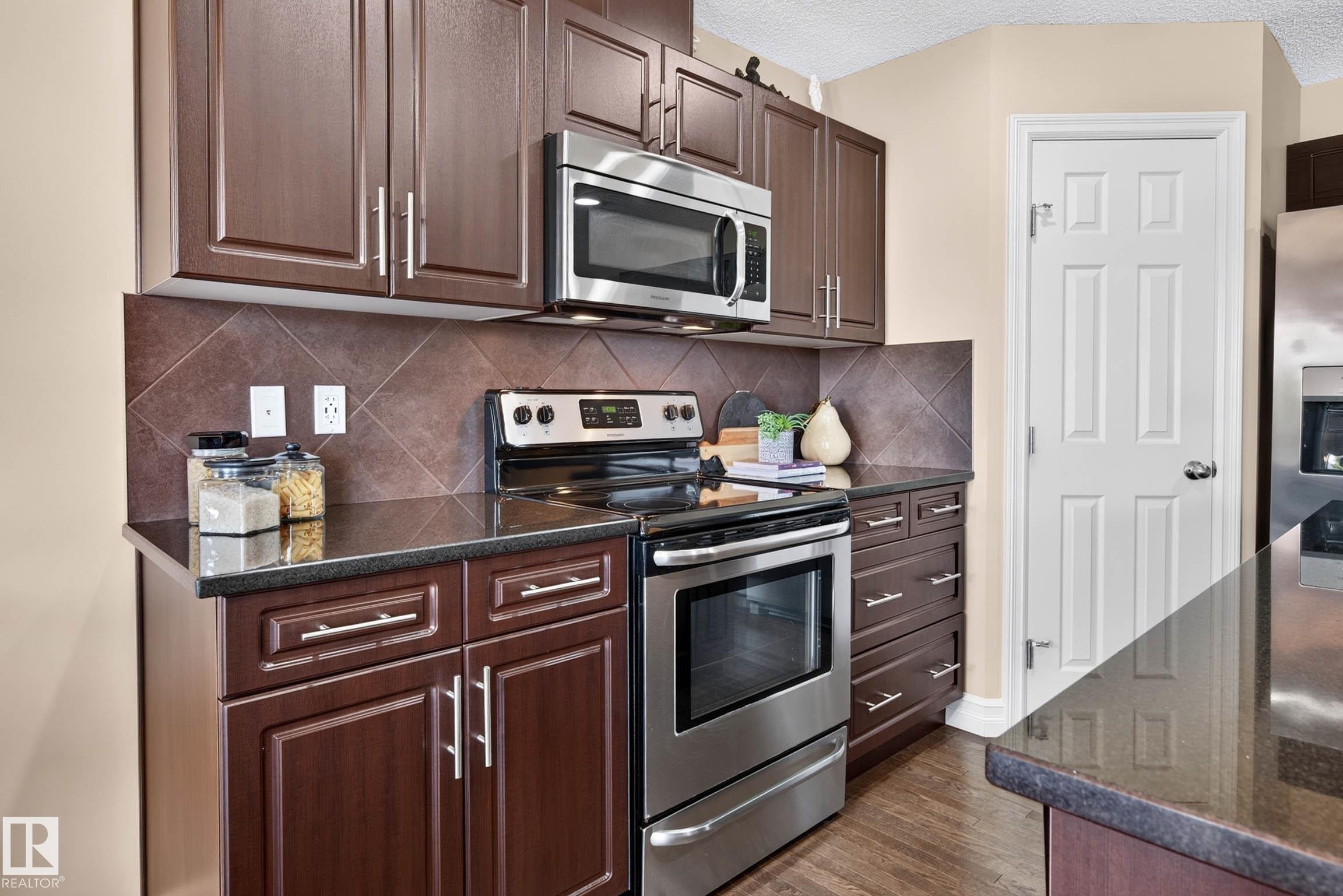 Kitchen featuring dark wood-finish cabinetry with silver-tone hardware, stainless steel appliances, dark countertops, and a tiled backsplash - 12119 167A Avenue, Edmonton, AB