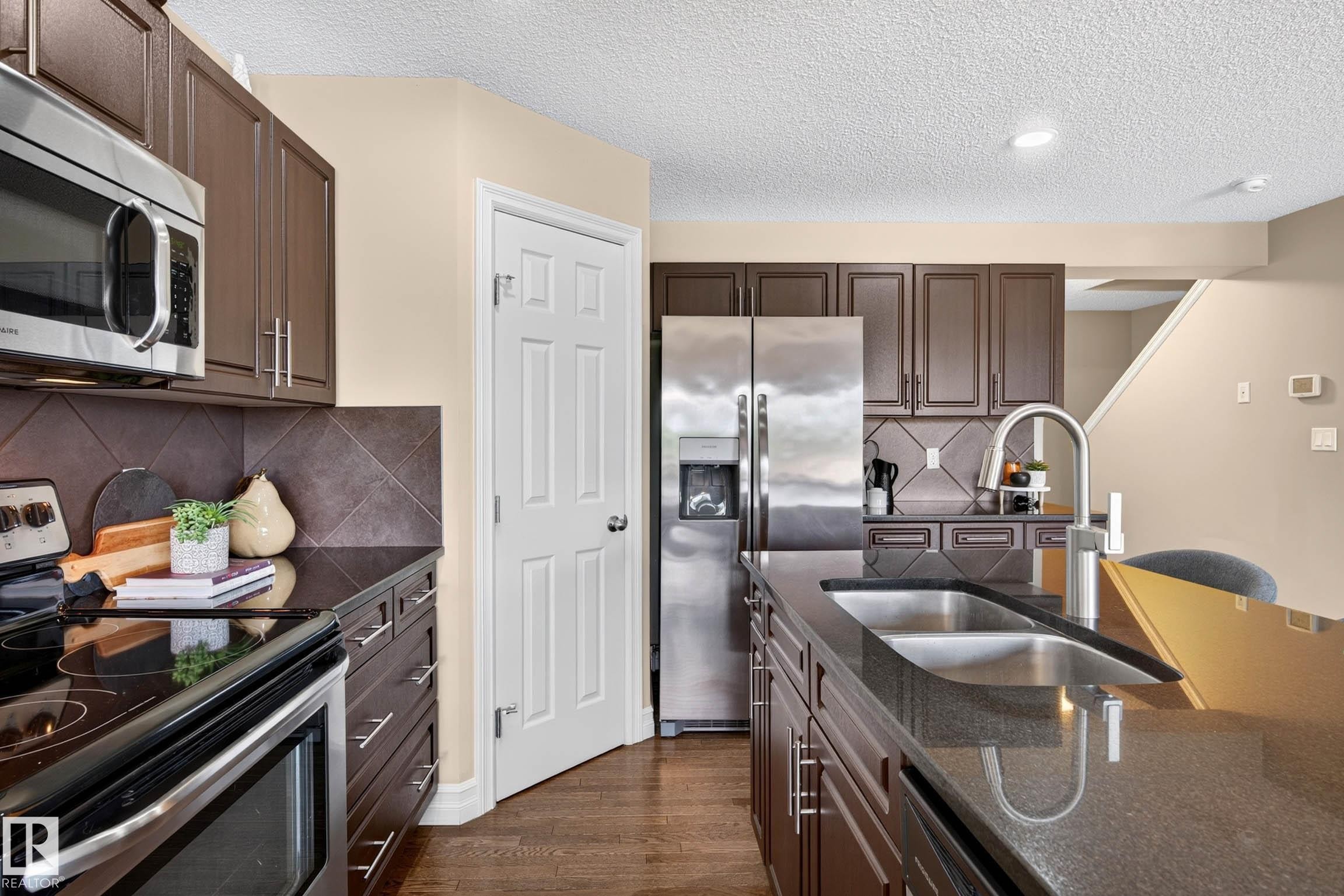 Kitchen featuring dark wood-finish cabinetry, stainless steel appliances, dark countertops, a tile backsplash, and a double basin sink with a gooseneck faucet - 12119 167A Avenue, Edmonton, AB