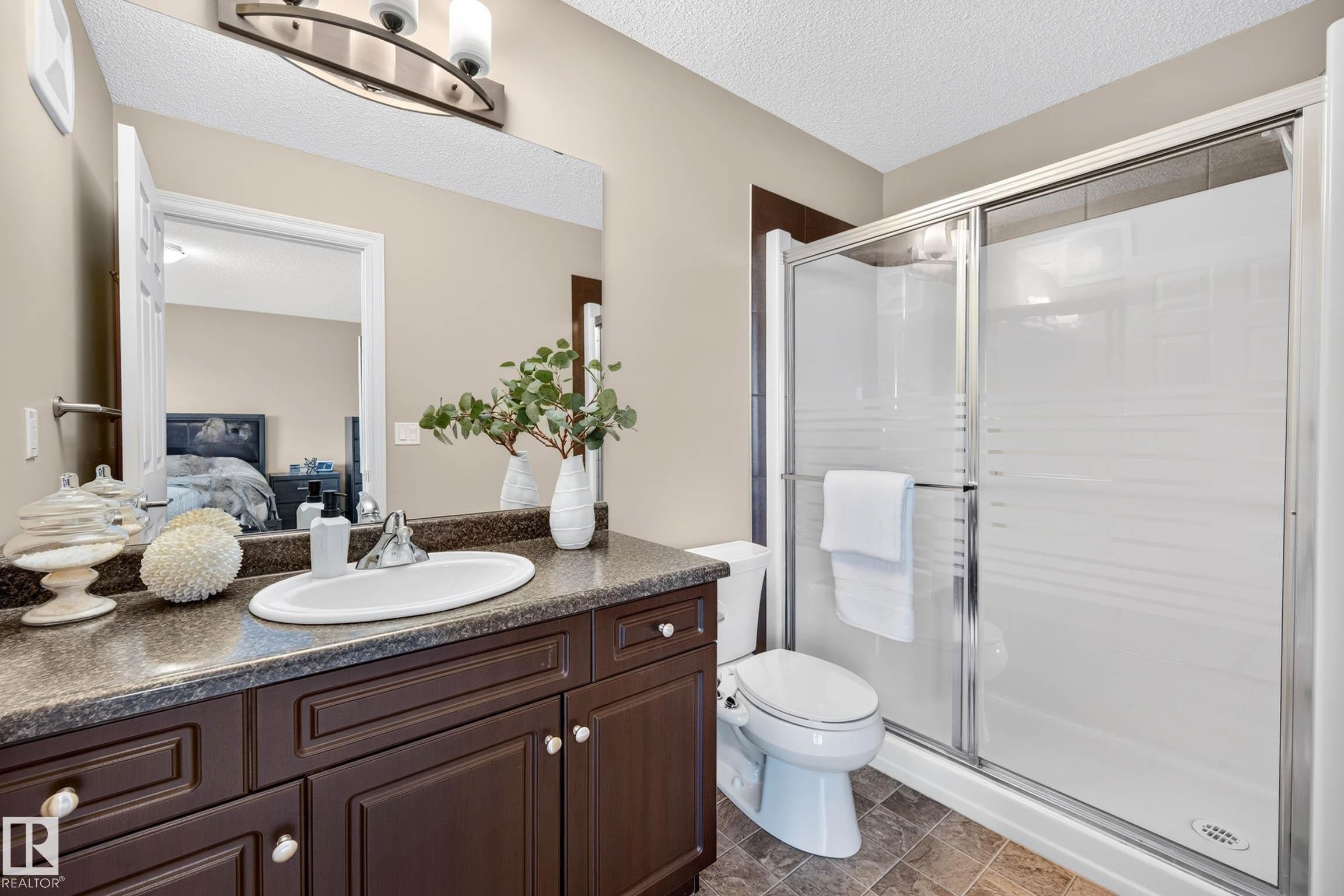 Bathroom featuring a dark wood-finish vanity with a built-in sink, a speckled countertop, a large wall-mounted mirror, and a glass-enclosed shower with frosted accents - 12119 167A Avenue, Edmonton, AB